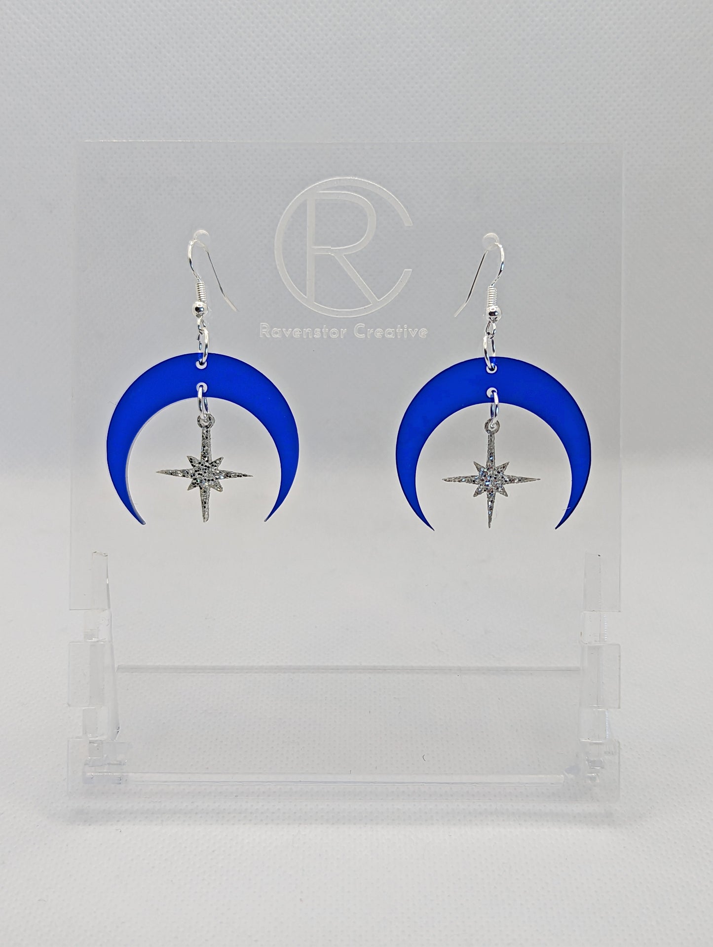 A pair of acrylic earrings that are two parts. Silver ear wire fixings are attached to the middle of a sideways crescent moon with a small star dangling in the centre. They have an Translucent Blue Glitter Moon with Silver Glitter star in the middle.