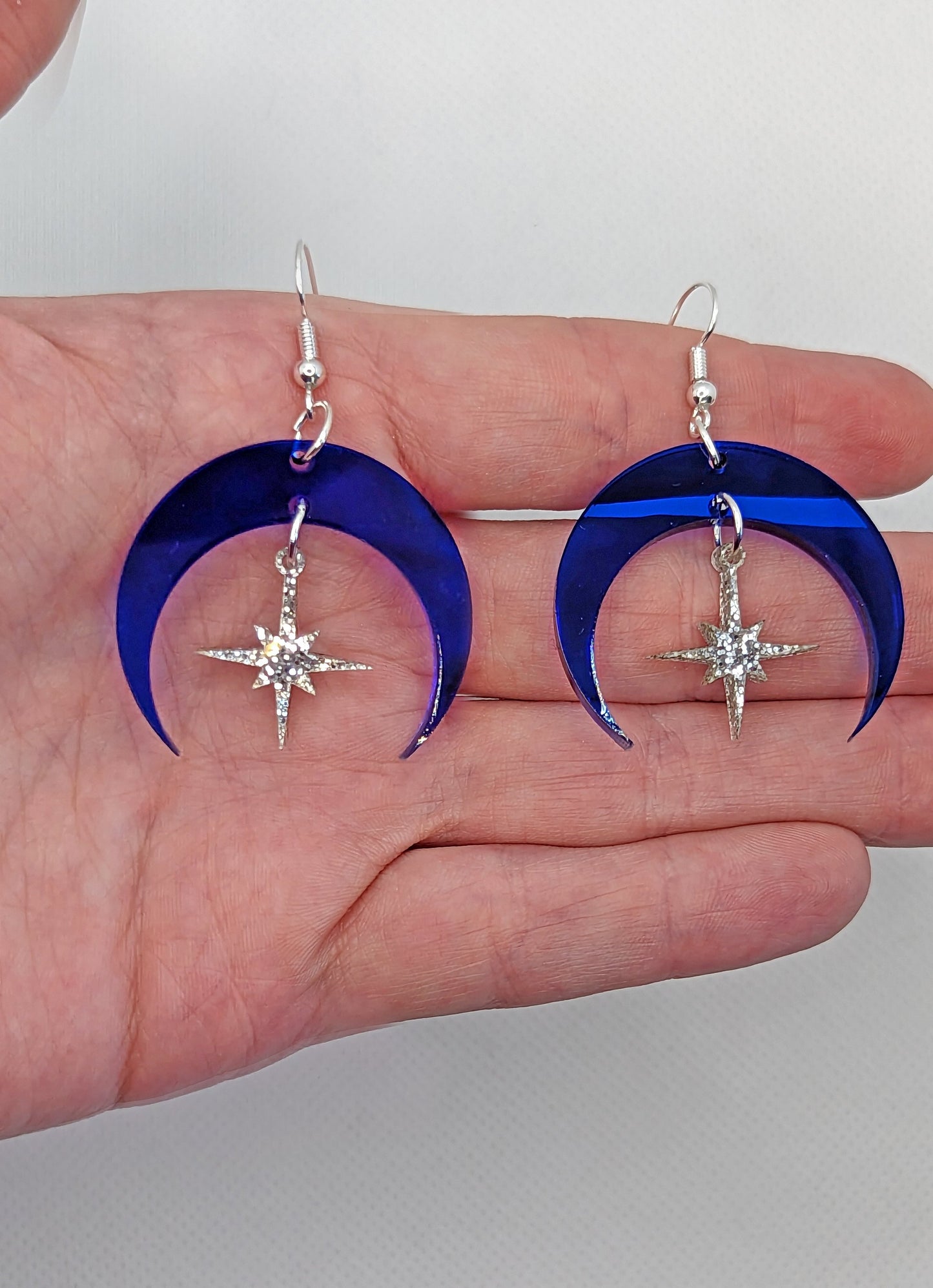 A pair of earrings that are two parts. Silver ear wire hook fixings are attached to a crescent moon shaped frame with a small star dangling in the middle. They are on a hand showing the size of three finger widths in length and two wide.