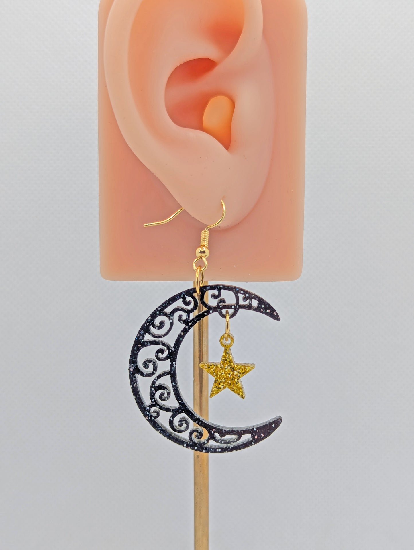 An acrylic earring with Gold ear wire hooks. It is a Crescent Moon that have an intricate pattern cut out with a star dangling in the middle. The Moon is Black and Silver Glitter and the star is Gold Glitter. It is on an ear to show the size.