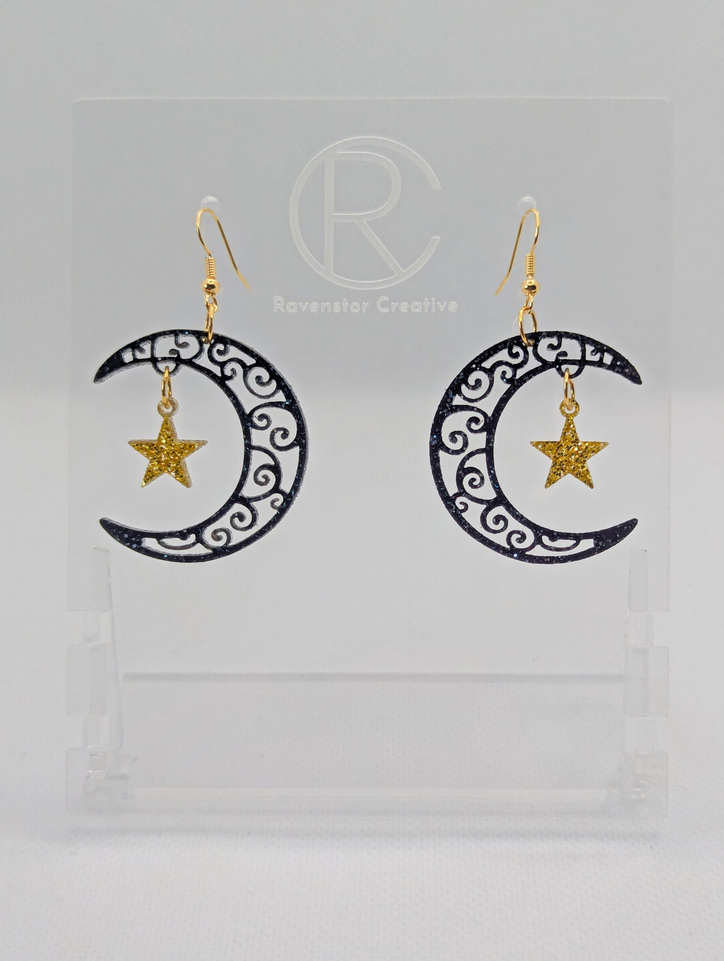 A pair of acrylic earrings with Gold ear wire hooks. They are Crescent Moons that have an intricate pattern cut out with a star dangling in the middle. The Moon is Black and Silver Glitter and the star is Gold Glitter.