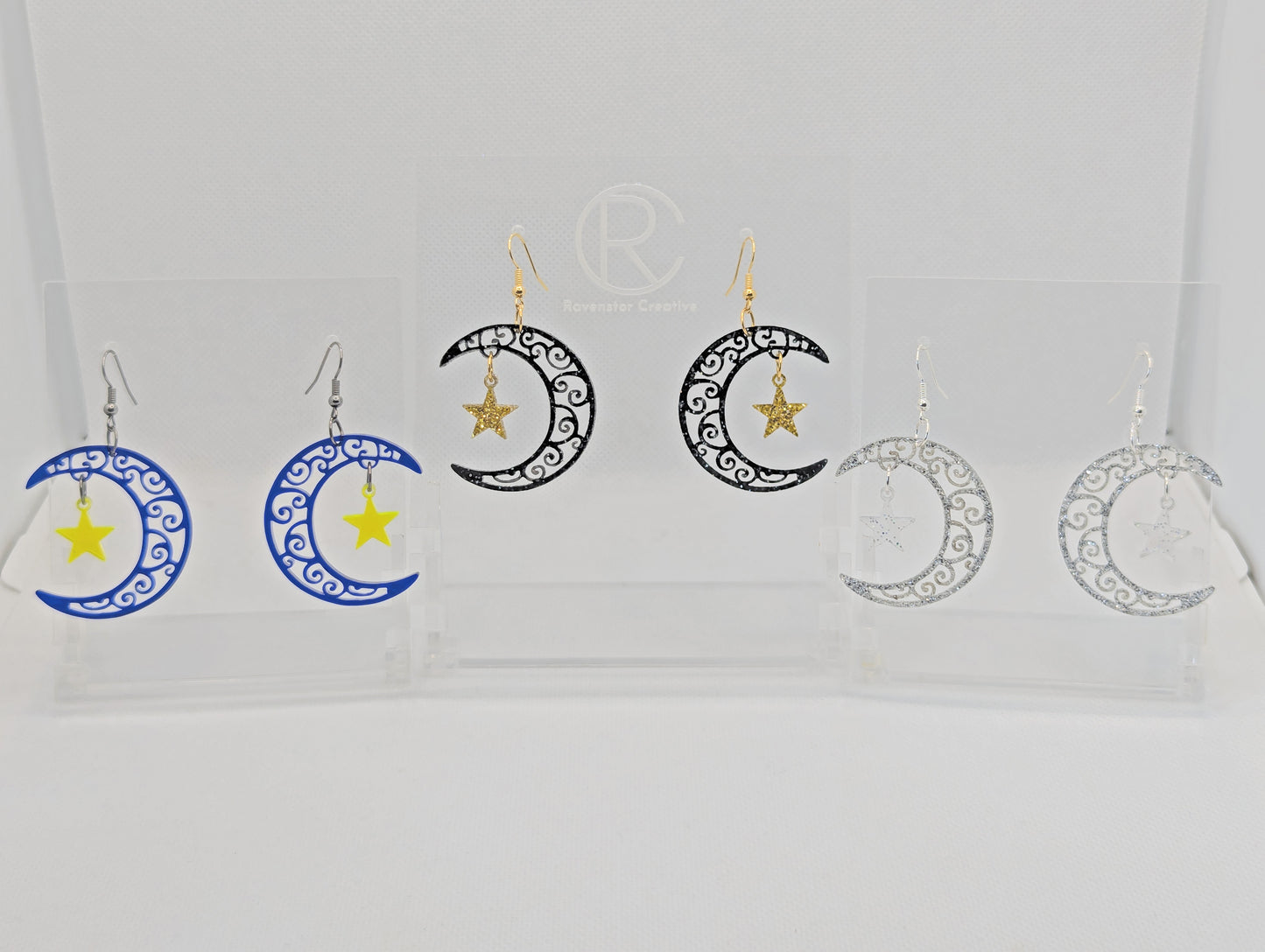 Three pairs of acrylic drop earrings. They are intricate Crescent Moons with a star dangling in the middle. The colours are Blue for the Moon and Neon Yellow Star. Black and Silver Glitter Moon and Gold glitter star. Silver Moon and Clear sparkle.