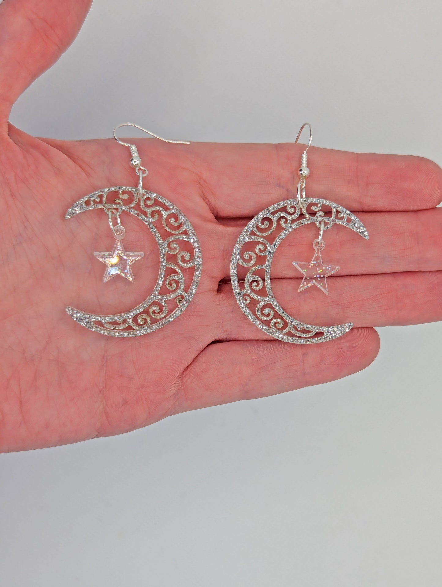 A pair of acrylic earrings with Silver ear wire hooks. They are Crescent Moons that have an intricate pattern cut out with a star dangling in the middle. They are on a hand to show the size of two and a half finger widths and the same in width.