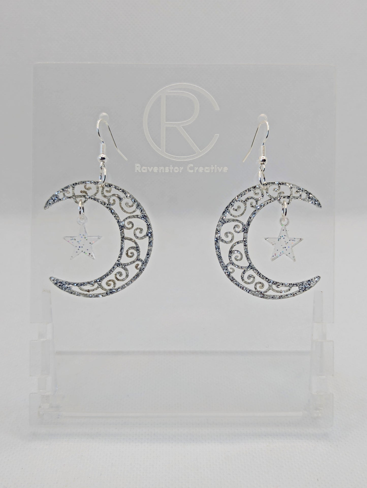A pair of acrylic earrings with Silver ear wire hooks. They are Crescent Moons that have an intricate pattern cut out with a star dangling in the middle. The Moon is Silver Glitter and the star is Clear Sparkle.