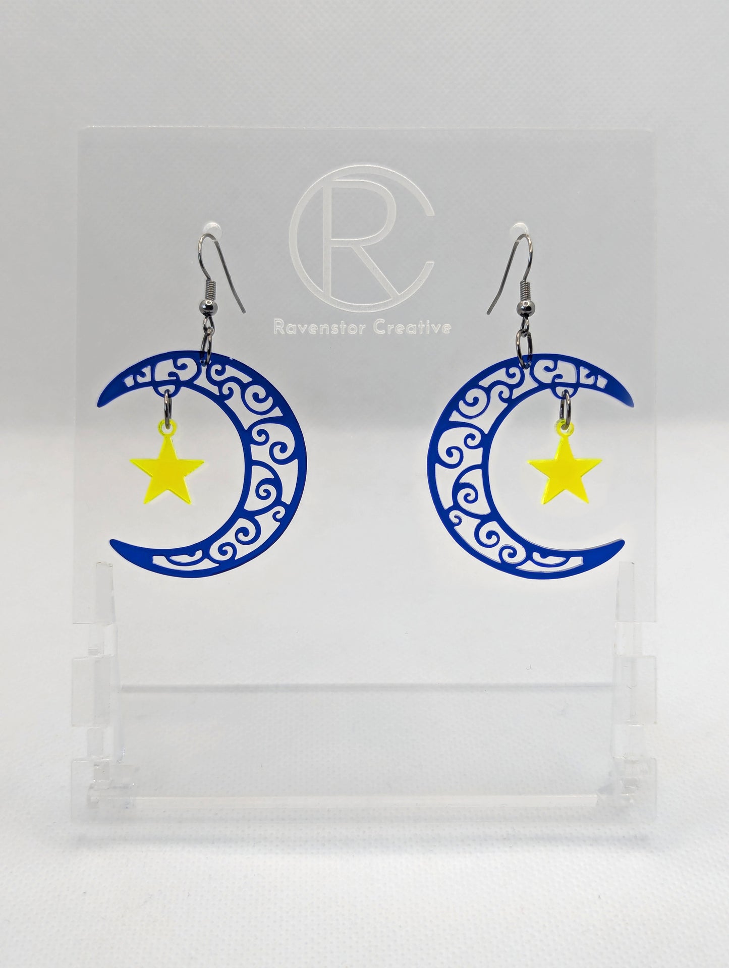 A pair of acrylic earrings with Black ear wire hooks. They are Crescent Moons that have an intricate pattern cut out with a star dangling in the middle. The Moon is Translucent Blue and the star is Neon Yellow.