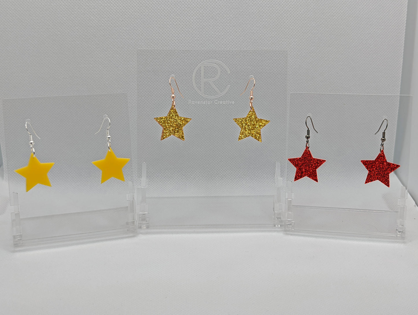 Shows three pairs of earrings that have ear wire hook fixings. They are five pointed star shaped. The colours are yellow, gold glitter and Red Glitter. They are against a white background on see through stand.
