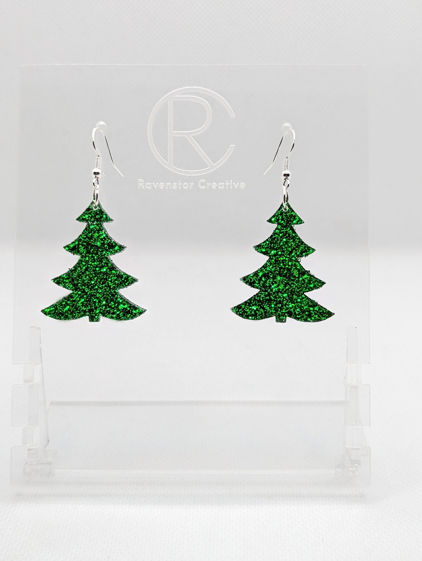 An image of a pair of earrings with Silver ear wire hook fixings. They are Christmas / Xmas Tree shapes. They are made in Green Glitter acrylic. They are dangling from a clear stand with the Ravenstor Creative logo on it against a white background