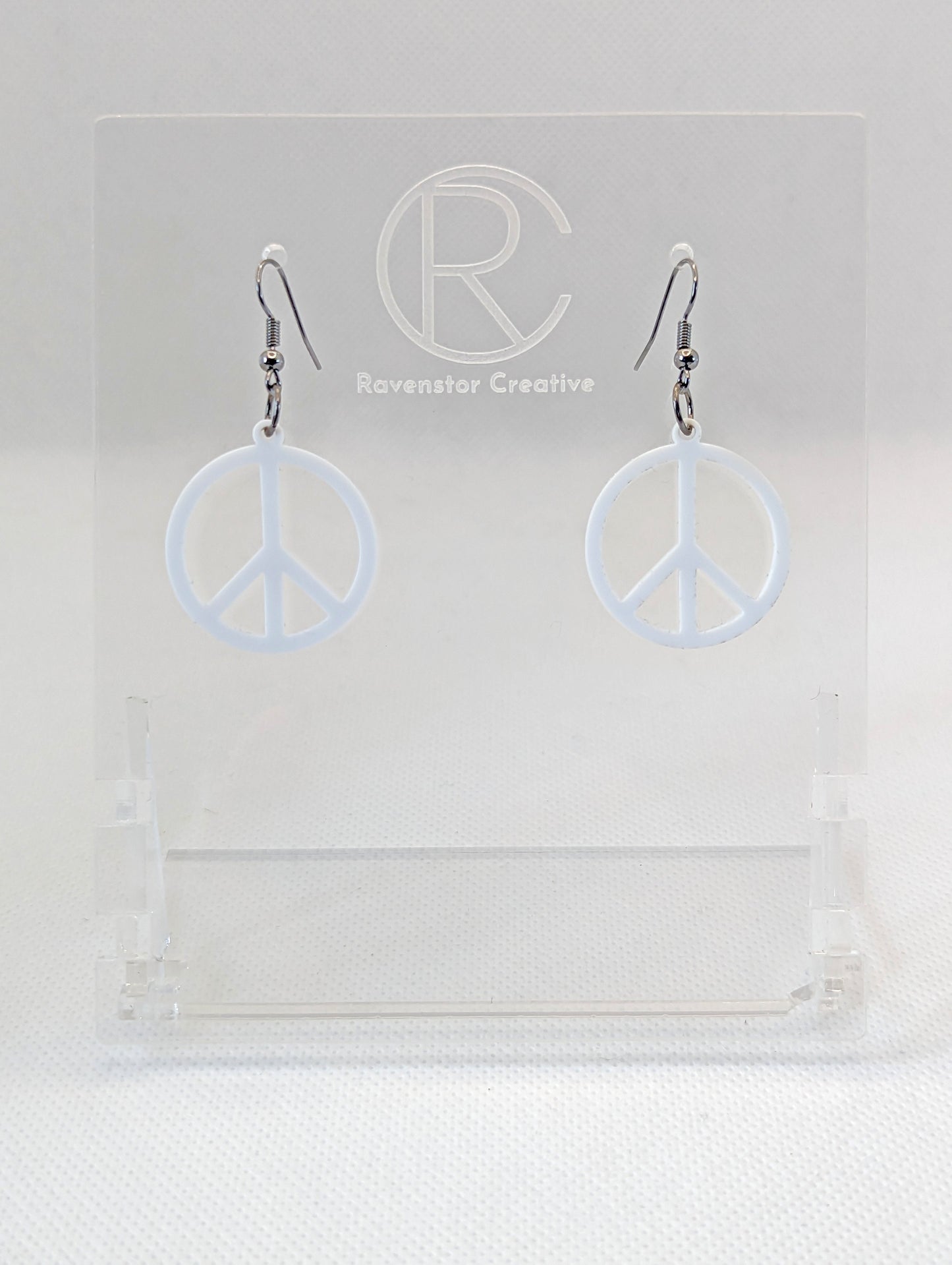 A pair of acrylic earrings in the shape of the 80s Retro Peace and love symbol. They have Black ear wire hook fixings and the colour is White. They are on a clear stand with the Ravenstor Creative Logo on it against a white background.