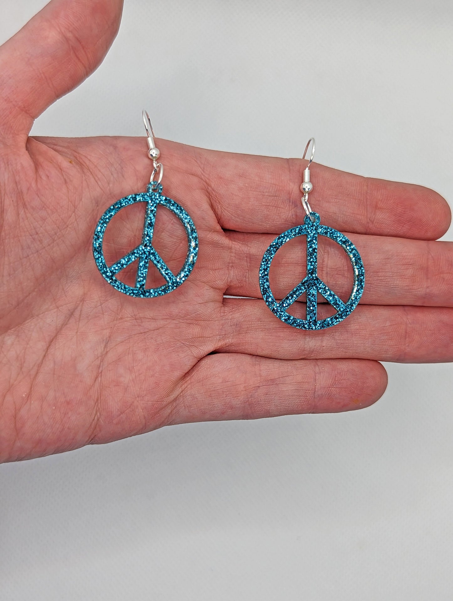 A pair of earrings in the shape of the 80s Retro Peace and love symbol. They have ear wire hook fixings and are in Ice Blue Glitter. They are against a hand to show the size of 2 finger widths length and 2 wide.