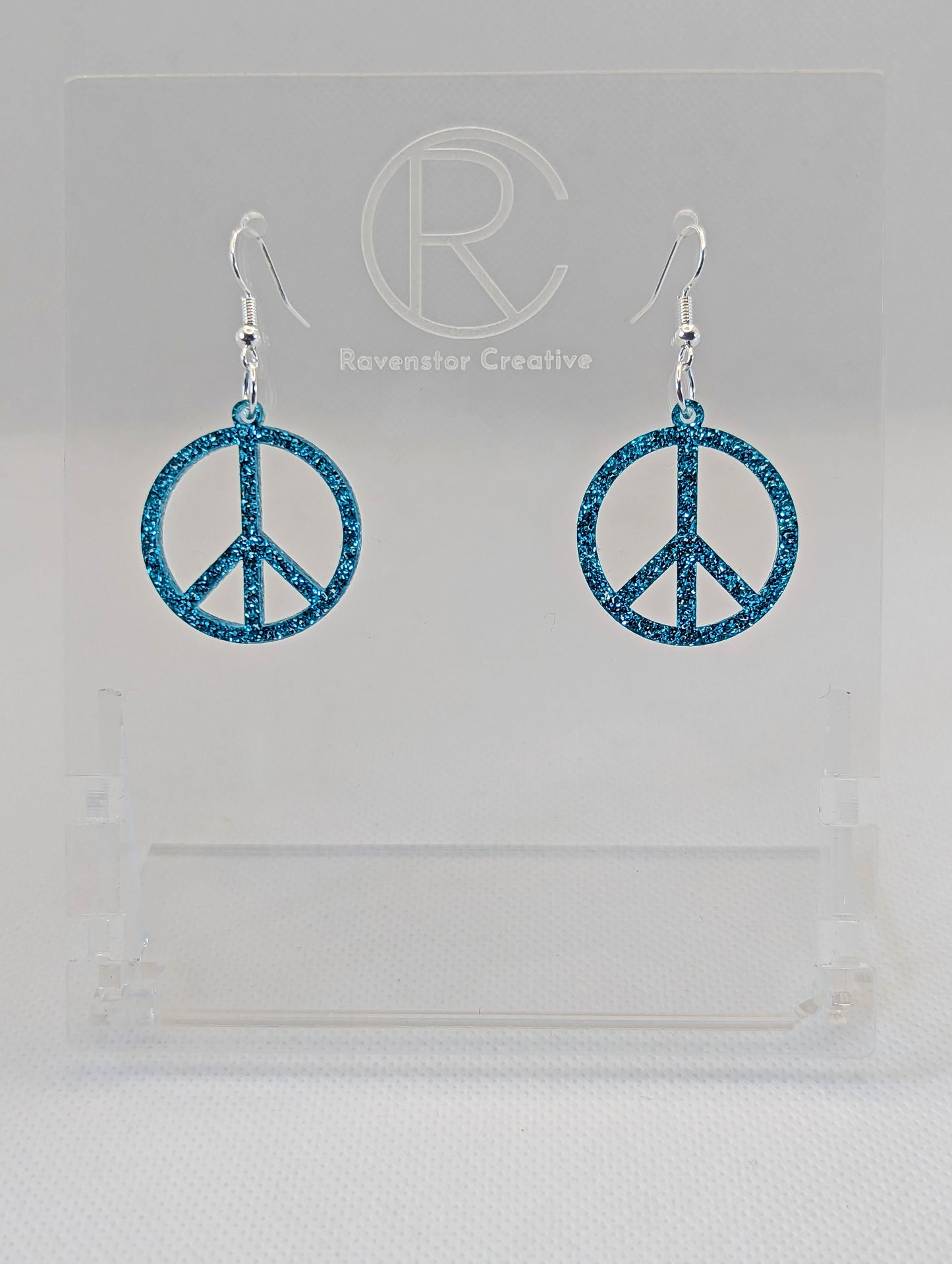 A pair of acrylic earrings in the shape of the 80s Retro Peace and love symbol. They have Silver ear wire hook fixings and the colour is Ice Blue Glitter. They are on a clear stand with the Ravenstor Creative Logo on it against a white background.
