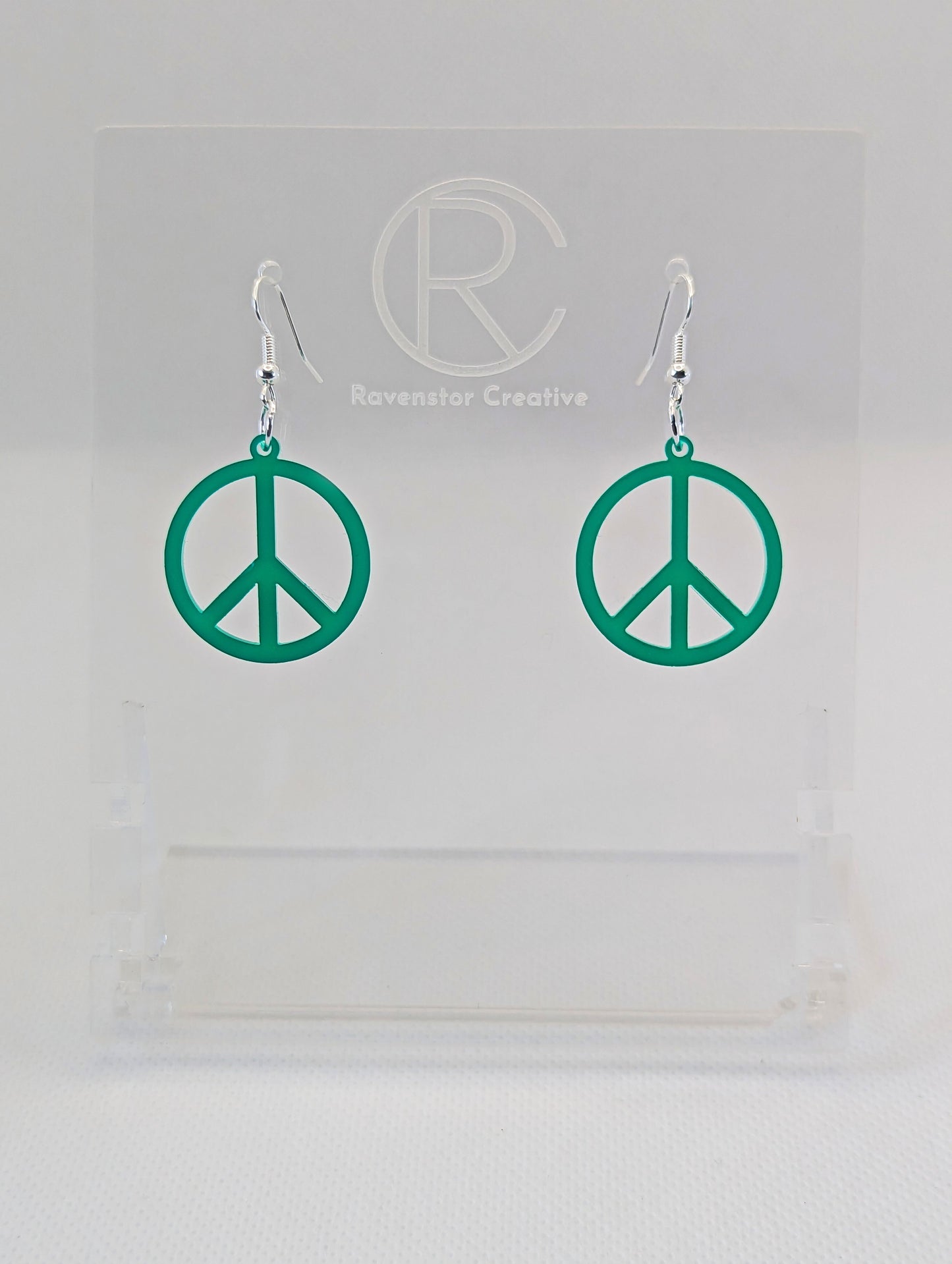 A pair of acrylic earrings in the shape of the 80s Retro Peace and love symbol. They have Silver ear wire hook fixings and the colour is Green. They are on a clear stand with the Ravenstor Creative Logo on it against a white background.