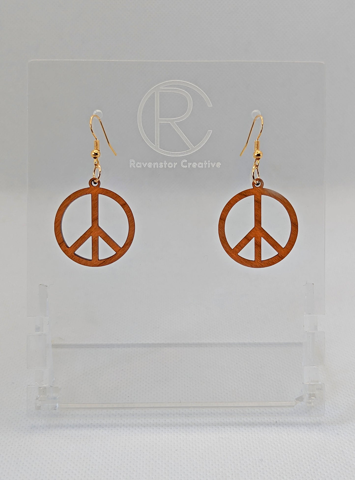 A pair of earrings in the shape of the 80s Retro Peace and love symbol. They have Gold ear wire hook fixings and they are Cherry Wood. They are on a clear stand with the Ravenstor Creative Logo on it against a white background.