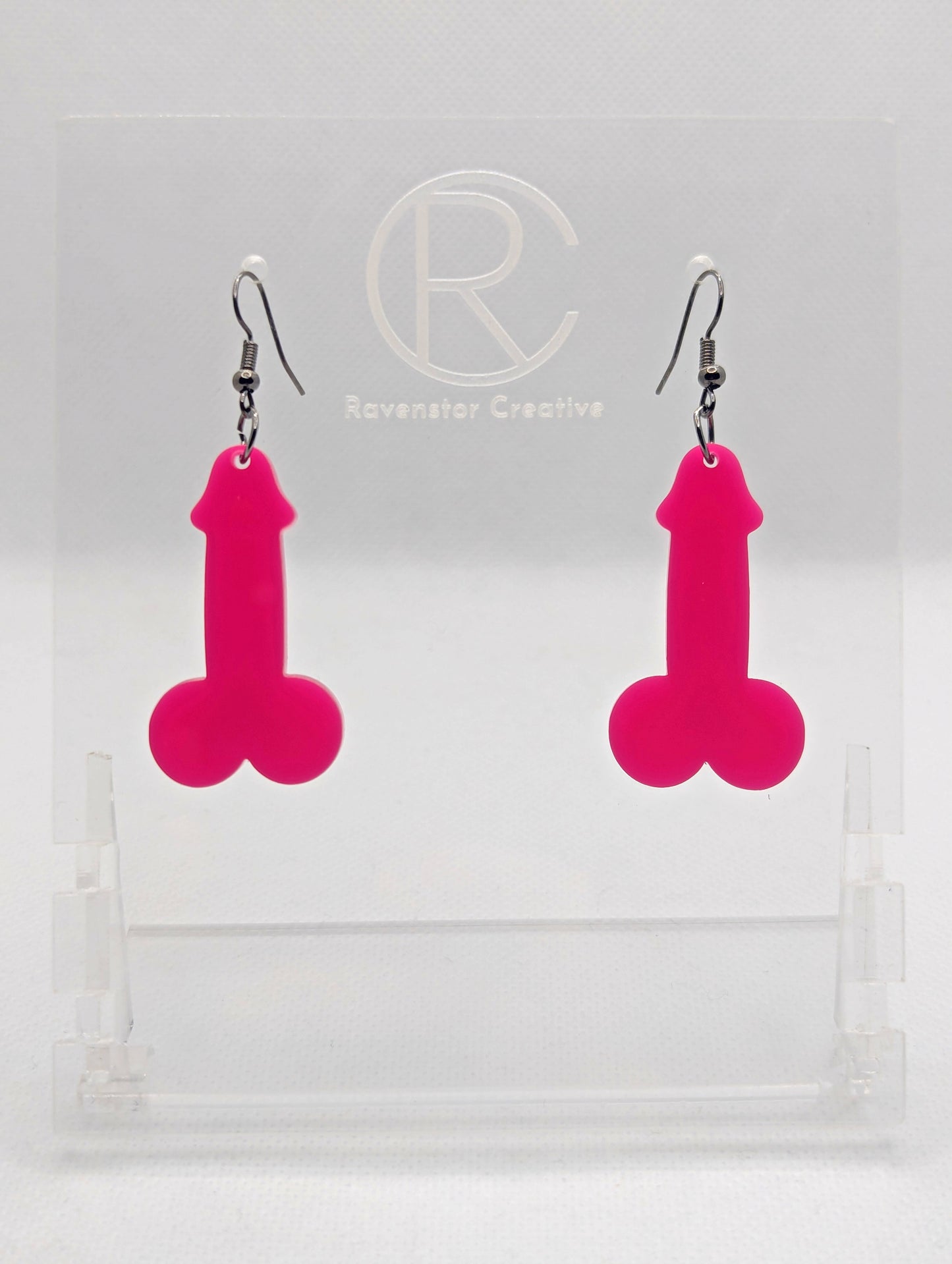 Image of a pair of earrings shaped like a Penis and Balls with Black wire hook fixings. These are ideal for funny Hen Dos or Bachelorette parties. The colour is Hot Pink and they are on clear stands against a white background.