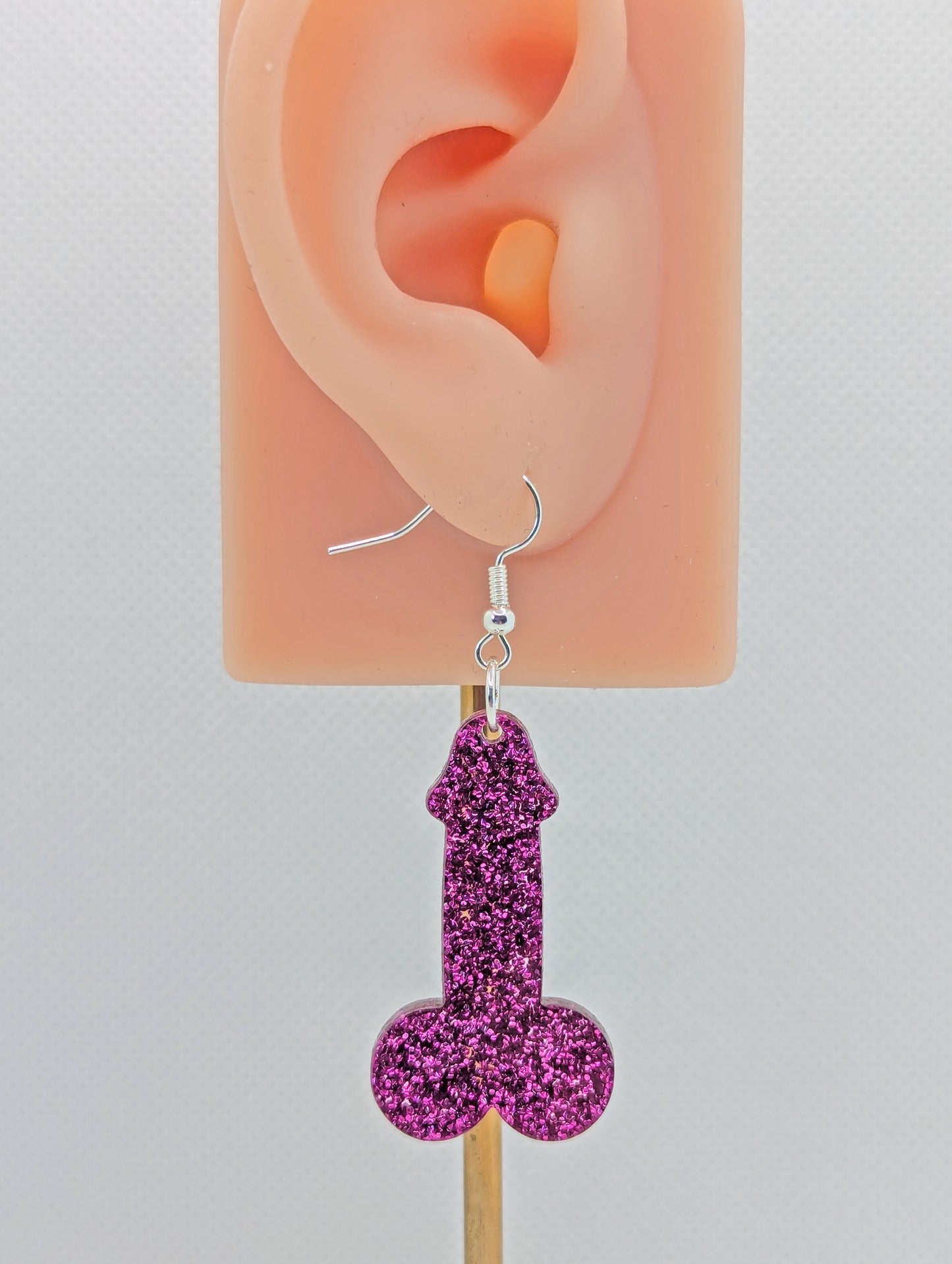 Image of an earring shaped like a Penis and Balls with a Silver ear wire hook fixing. These are ideal for funny Hen Dos or Bachelorette parties. The colour is Pink Glitter and is on an ear to show the size.