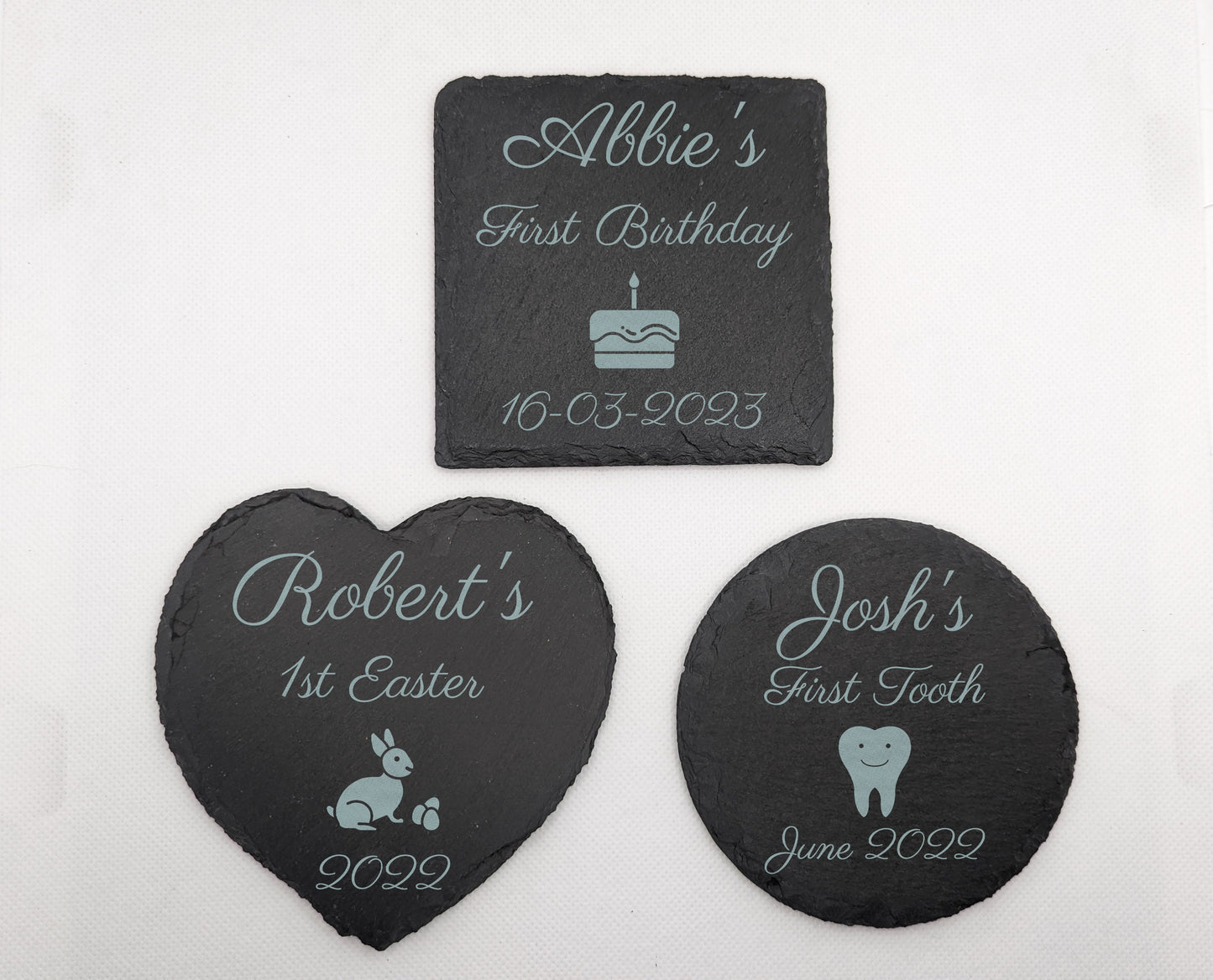 3 slate coasters engraved with Babys first themes. The Square has Abbies First Birthday with a cake and date. The heart has Roberts 1st Easter with abunny and year. The circle has Joshs First Tooth with a cartoon tooth and Date.