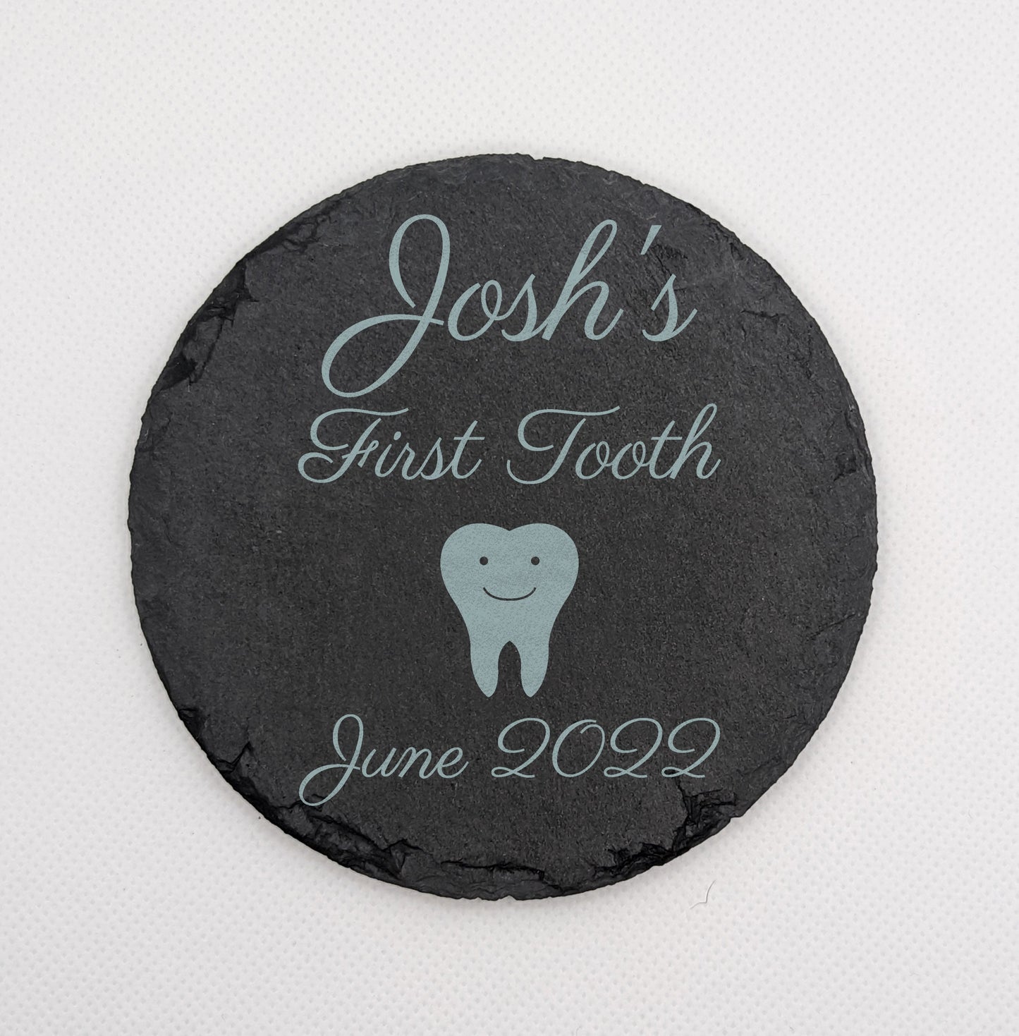Shows a babys first themed slate circle coaster 10cm by 10cm. It is engraved with example text saying Joshs First Tooth June 2022 with the image of a cartoon tooth with smiley face. It is stand against a white background.