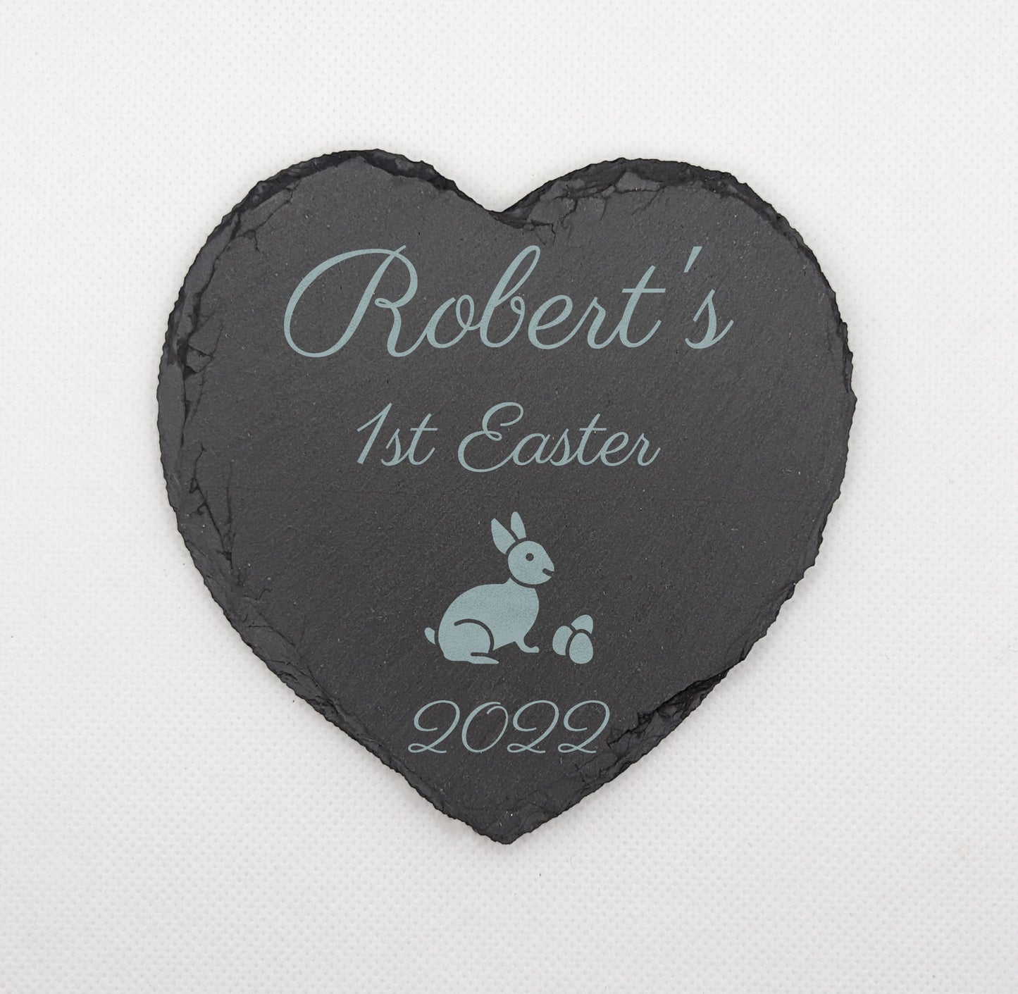 Shows a babys first themed Slate Heart Coaster 10cm by 10cm. It is engraved with example text saying Roberts 1st Easter 2022 with the image of an Easter bunny. It is on against a white background.