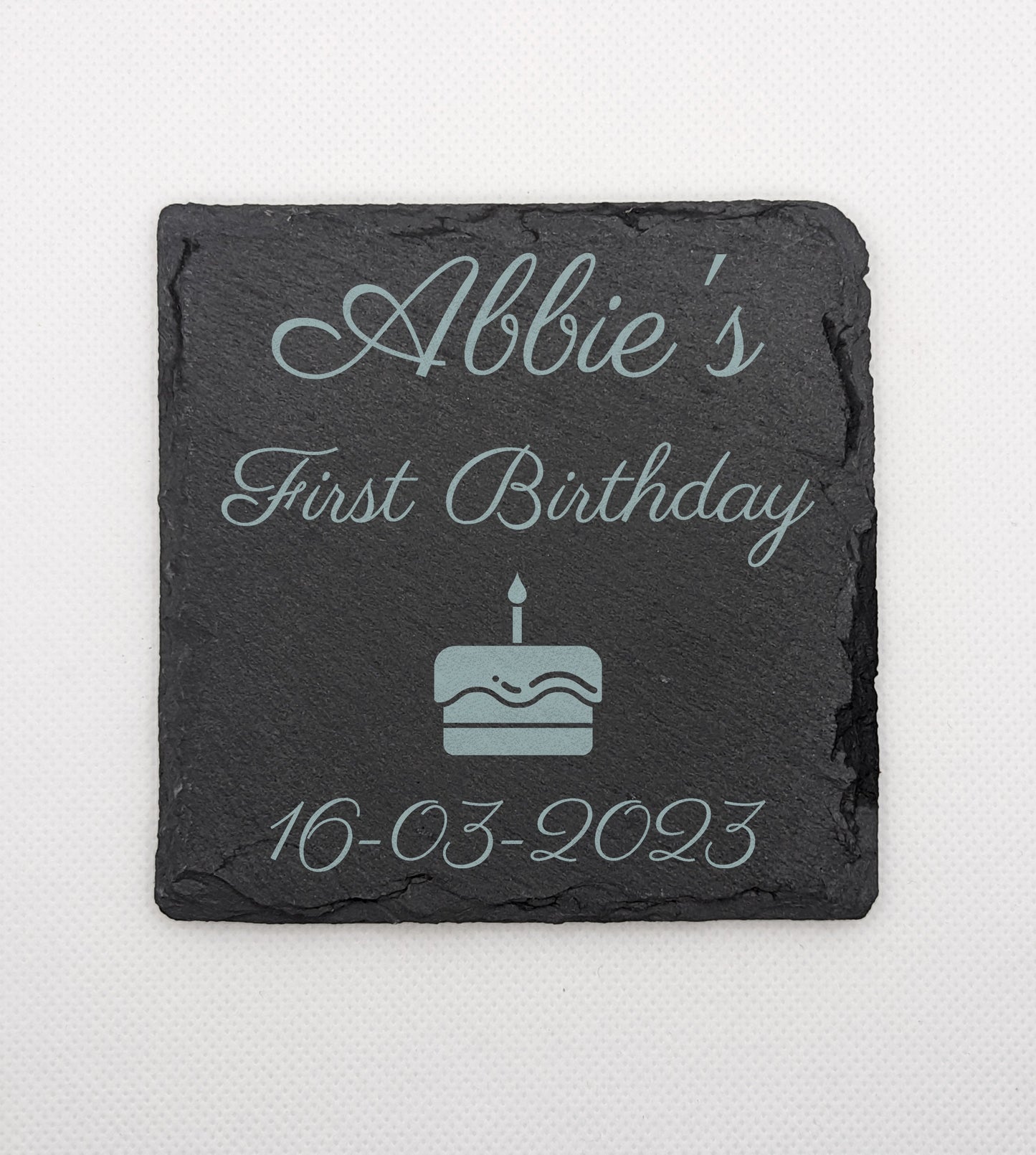 Shows a babys first themed square slate coaster 10cm by 10cm. It is engraved with example text saying Abbies First Birthday 16.03.2023 with an image of a Birthday cake with one candle. It is against a white background.