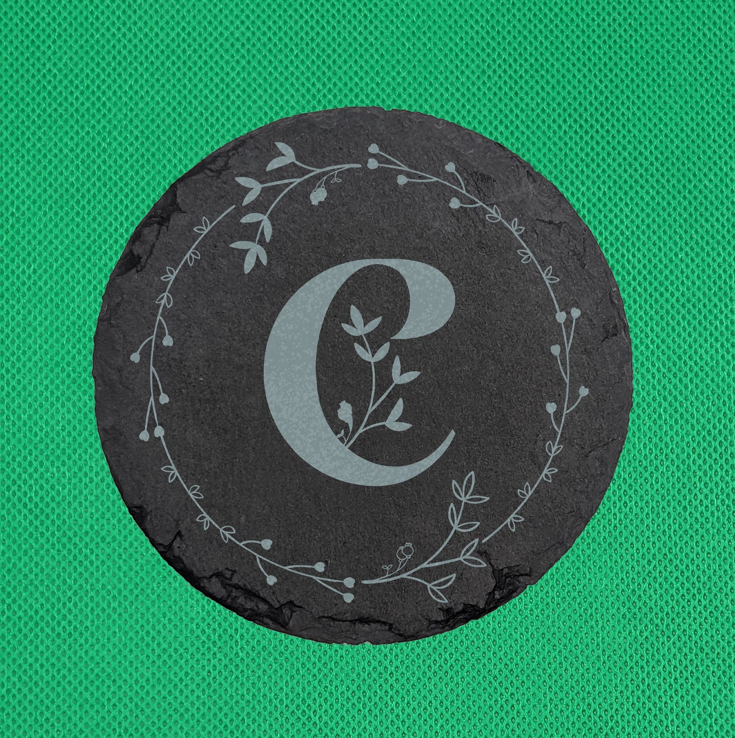 Shows a circle slate coaster engraved with the example initial C in a Bold font surrounded by a wreath of leaves and tiny hearts. It is on a clear stand against a red background. It is against a Green Background.