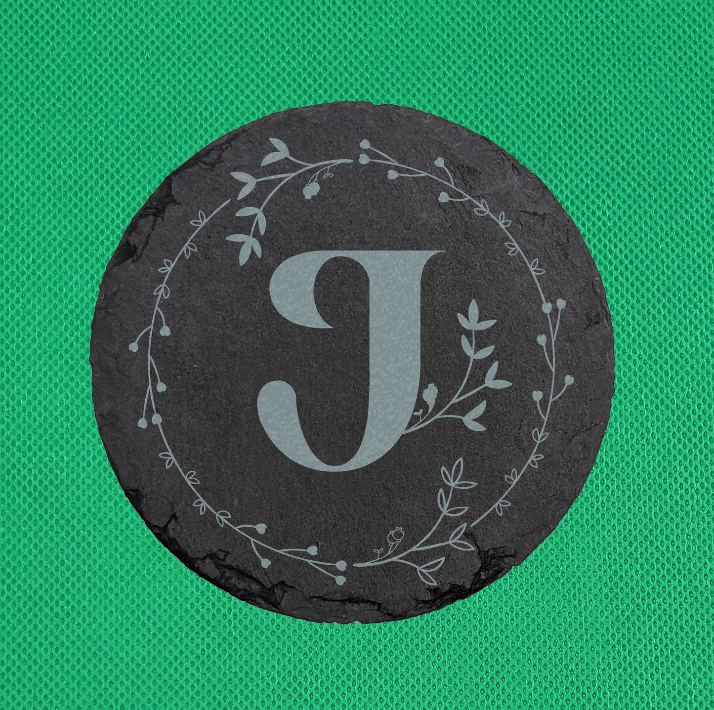 Shows a circle slate coaster engraved with the example initial J in a Bold font surrounded by a wreath of leaves and tiny hearts. It is on a clear stand against a red background. It is against a Green Background.