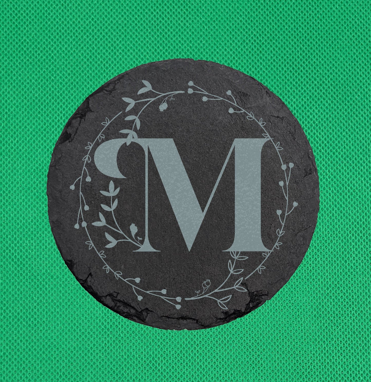 Shows a circle slate coaster engraved with the example initial M in a Bold font surrounded by a wreath of leaves and tiny hearts. It is on a clear stand against a red background. It is against a Green Background.