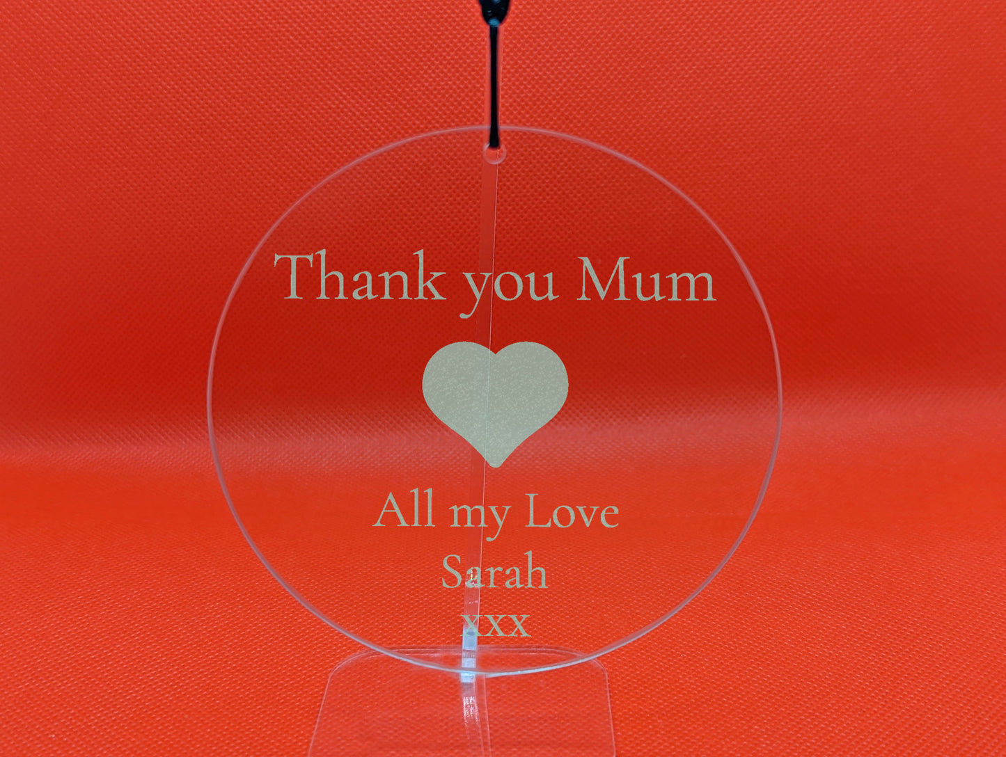Shows a mum themed clear hanging glass circle 10cm by 10cm. It is engraved with example text saying Thank you Mum All my Love Sarah xxx with an image a Heart in the middle. It is against a Red background on a clear stand.