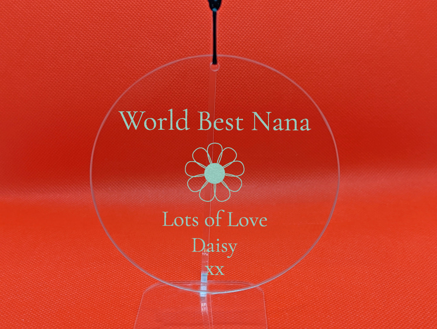 Shows a mum themed clear hanging glass circle 10cm by 10cm. It is engraved with example text saying Worlds Best Nana Lots of Love Daisy xx with the image of a Daisy Flower in the middle. It is against a Red background on a clear stand.