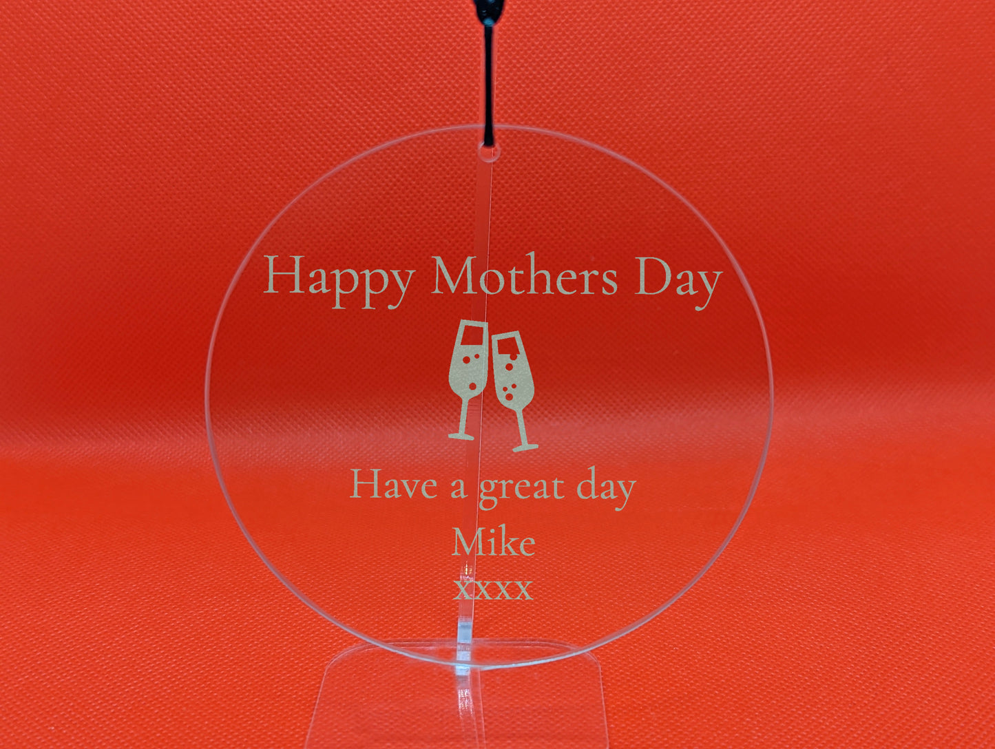 Shows a mum themed clear hanging glass circle 10cm by 10cm. It is engraved with example text saying Happy Mothers Day Have a great day Mike xxxx with an image of Two Champaign Glasses in the middle. It is against a Red background on a clear stand.