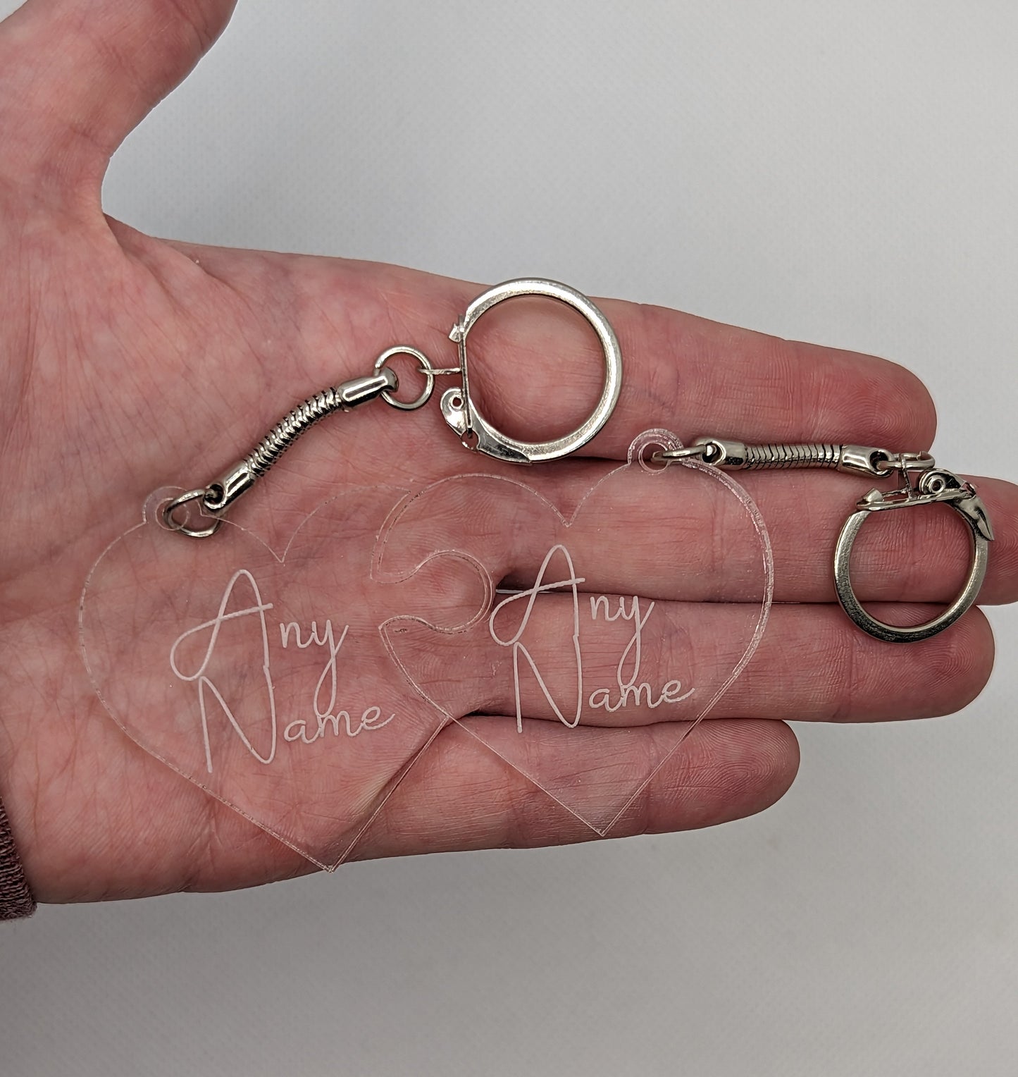 A Pair of interlocking heart keyrings, linked together like jigsaw pieces. They are made from clear acrylic and have the example engraving of Any Name on them. They are against a hand to show the size of 3 finger widths in length and width each
