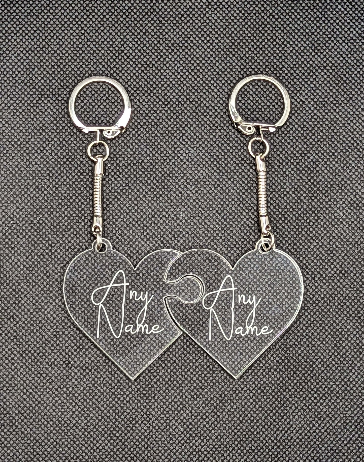 A Pair of interlocking heart keyrings / keychains, linked together like jigsaw pieces. They are made from clear / see through acrylic and have the example engraving of Any Name on them. They are against a dark background.