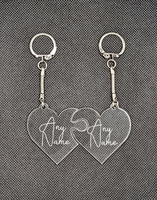 A Pair of interlocking heart keyrings / keychains, linked together like jigsaw pieces. They are made from clear / see through acrylic and have the example engraving of Any Name on them. They are against a dark background.