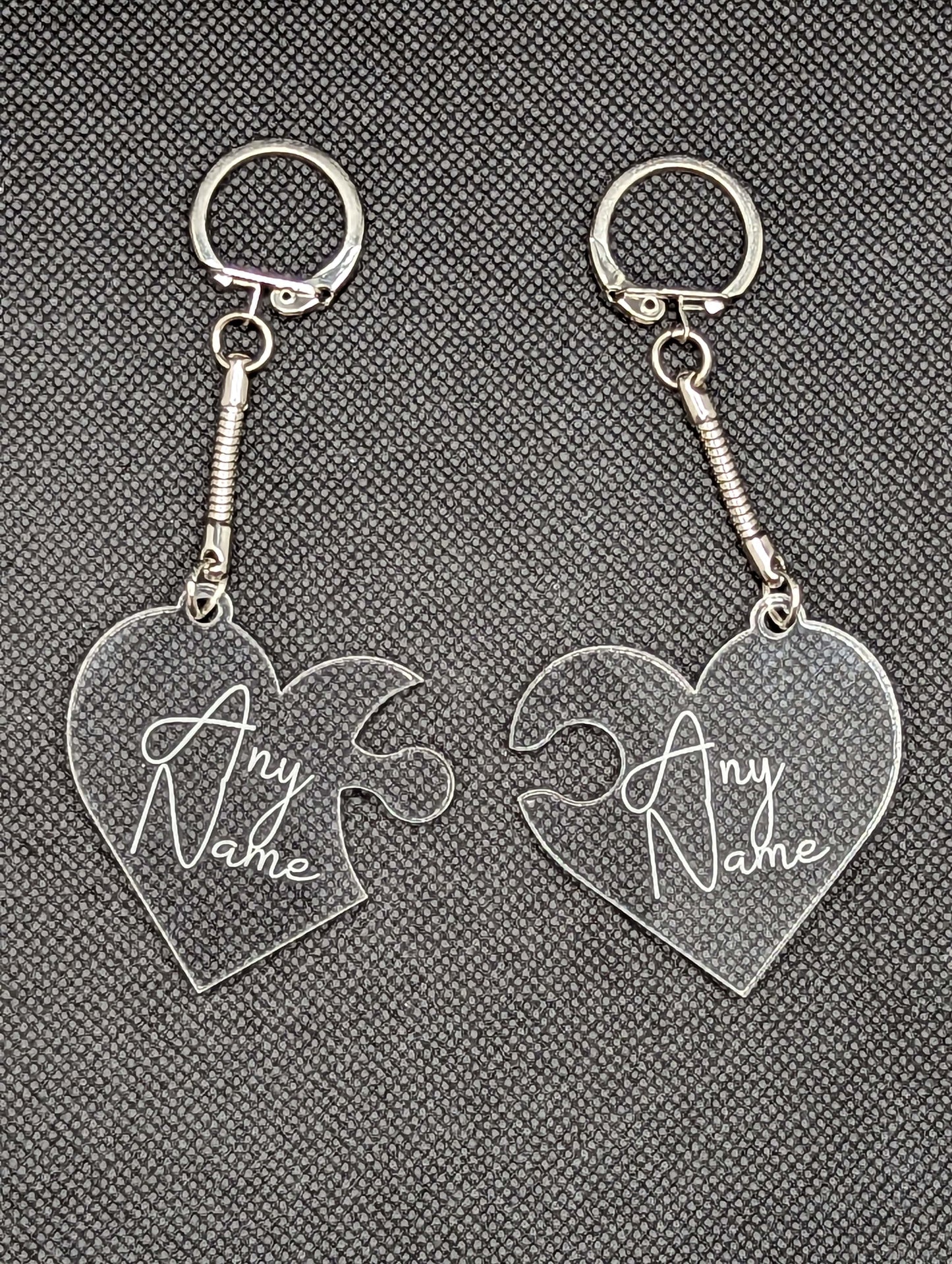 A Pair of interlocking heart keyrings / keychains, linked together like jigsaw pieces. They are made from clear / see through acrylic and have the example engraving of Any Name on them. They are against a dark background.