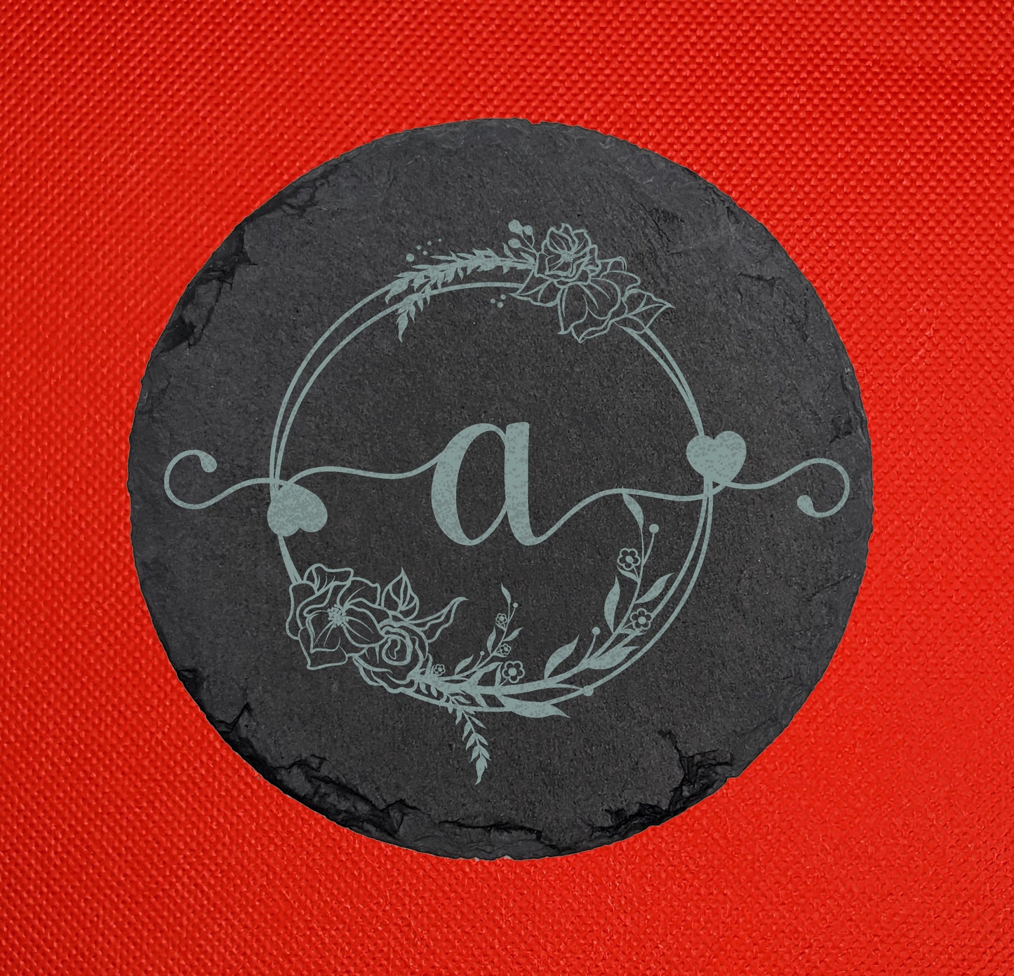 Shows a slate circle coaster engraved with the example initial Lower case a in a fancy font with hearts coming out from it. It is surrounded by a wreath with flowers and leaves on it. It is against a red background.