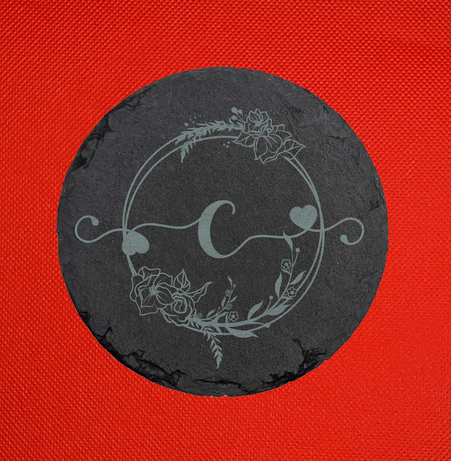 Shows a slate circle coaster engraved with the example initial Lower case c in a fancy font with hearts coming out from it. It is surrounded by a wreath with flowers and leaves on it. It is against a red background.