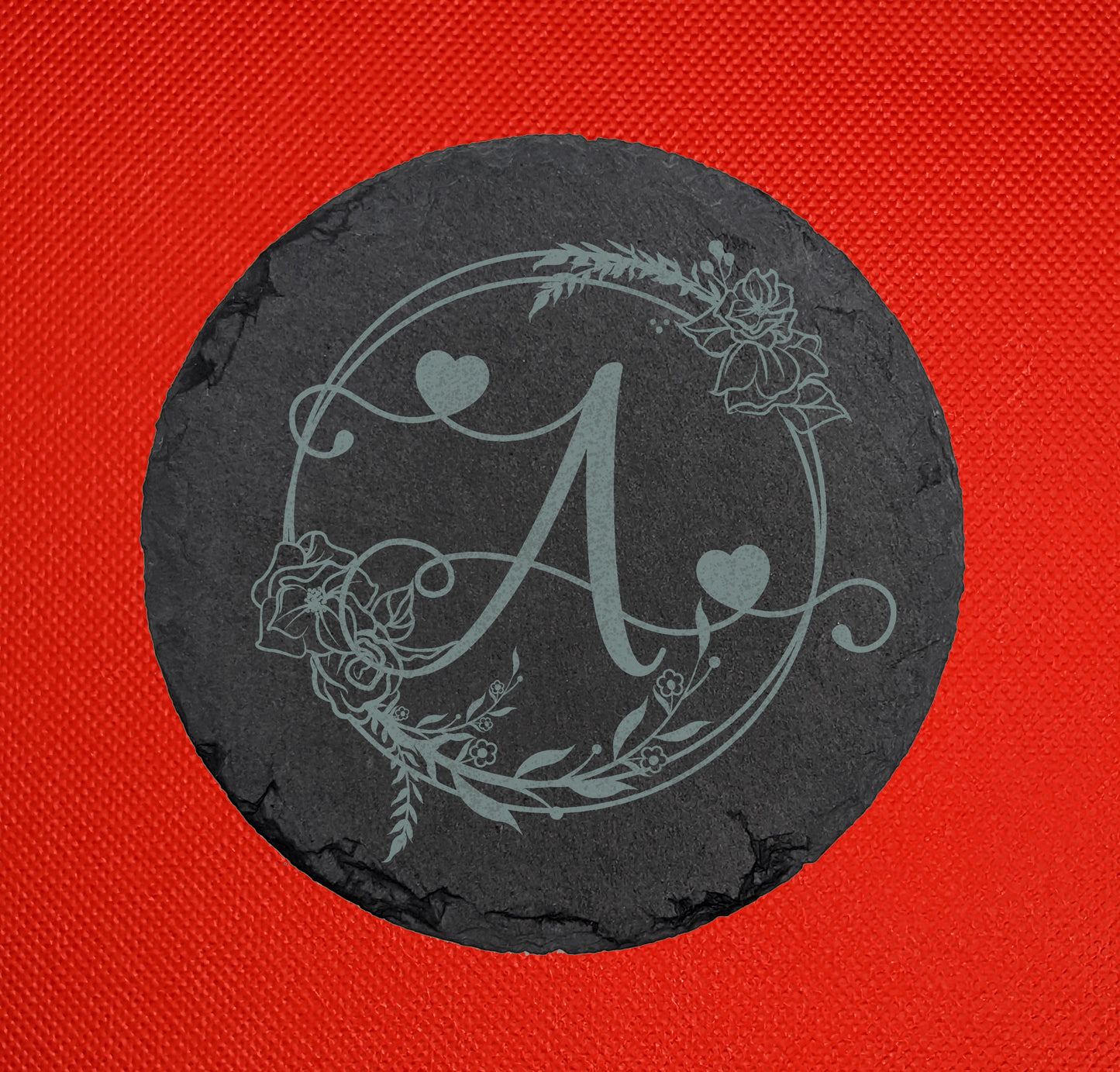 Shows a slate circle coaster engraved with the example initial Capital / Upper case A in a fancy font with hearts coming out from it. It is surrounded by a wreath with flowers and leaves on it. It is against a red background.