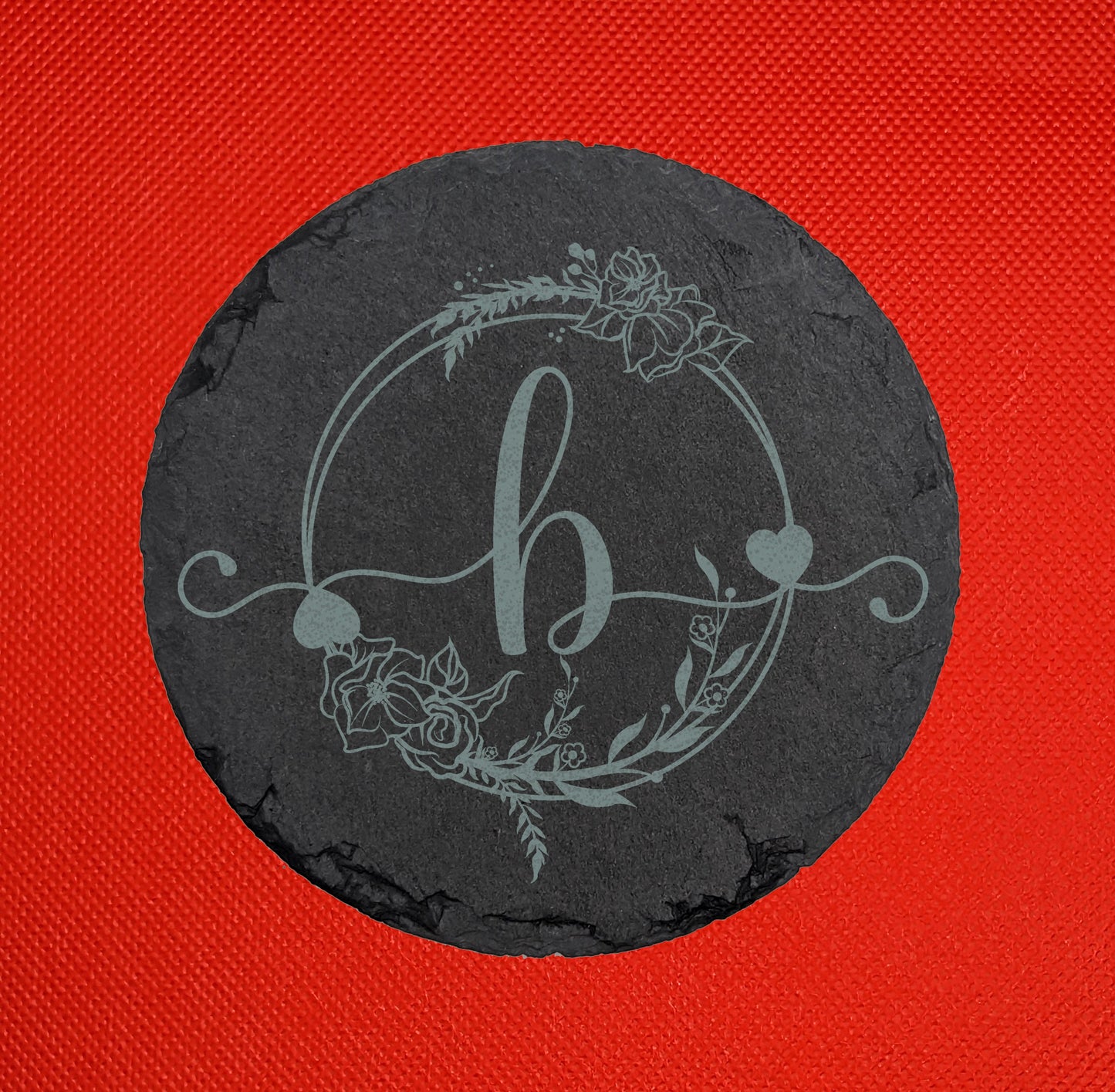 Shows a circle slate coaster engraved with the example initial Lower case b in a fancy font with hearts coming out from it. It is surrounded by a wreath with flowers and leaves on it. It is against a red background.