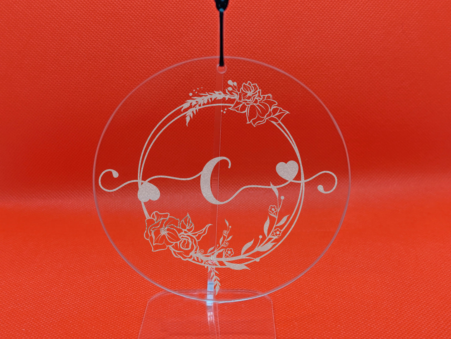 Shows a hanging glass circle engraved with the example initial Lower case c in a fancy font with hearts coming out from it. It is surrounded by a wreath with flowers and leaves on it. It is on a clear stand against a red background.