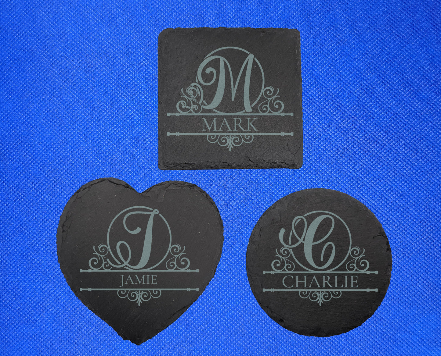 Three example Sale Coasters. They are engraved with initial letter in an elegant font with fancy patterns around it. Under is a name between two bars. The square has an M with Mark under, Heart has J with Jamie under, Circle has C with Carlie under.