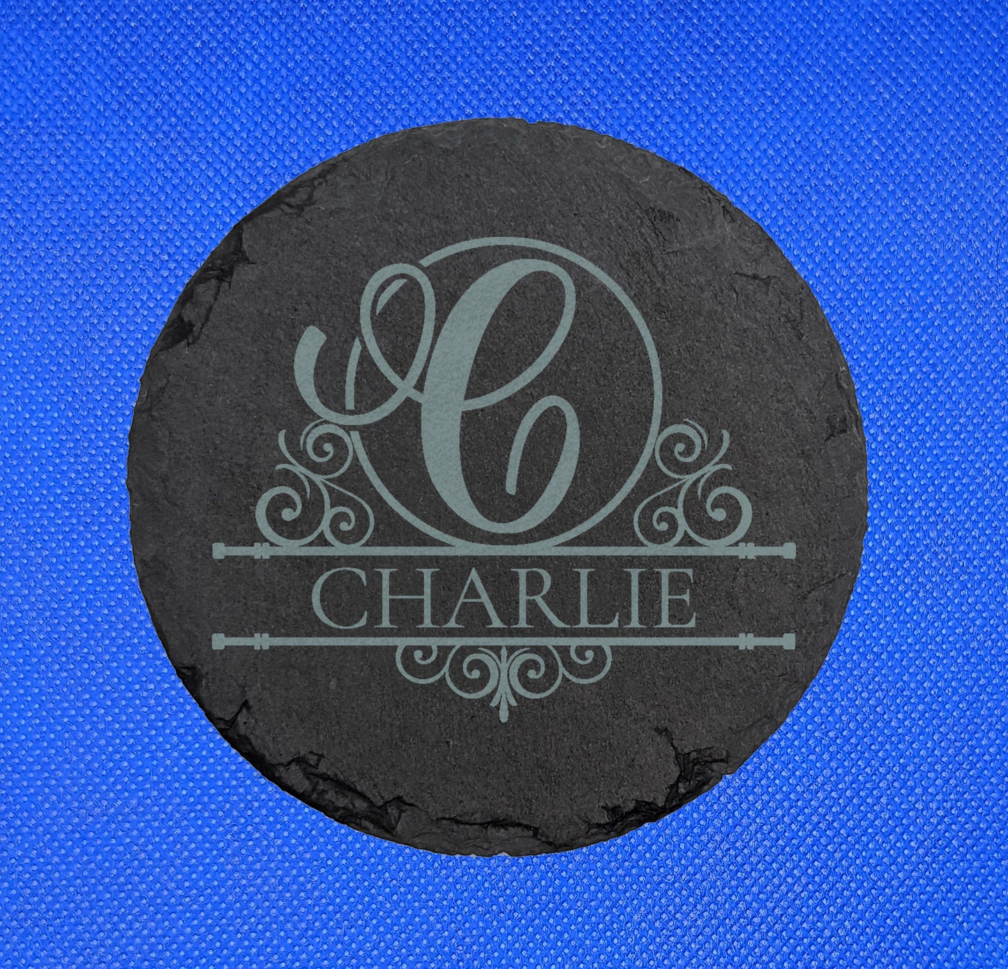 Shows a Slate Circle Coaster engraved with the example Initial C in elegant lettering and fancy patterns around it. Under is the name CHARLIE between two horizontal bars with more patterns. It is against a blue background.