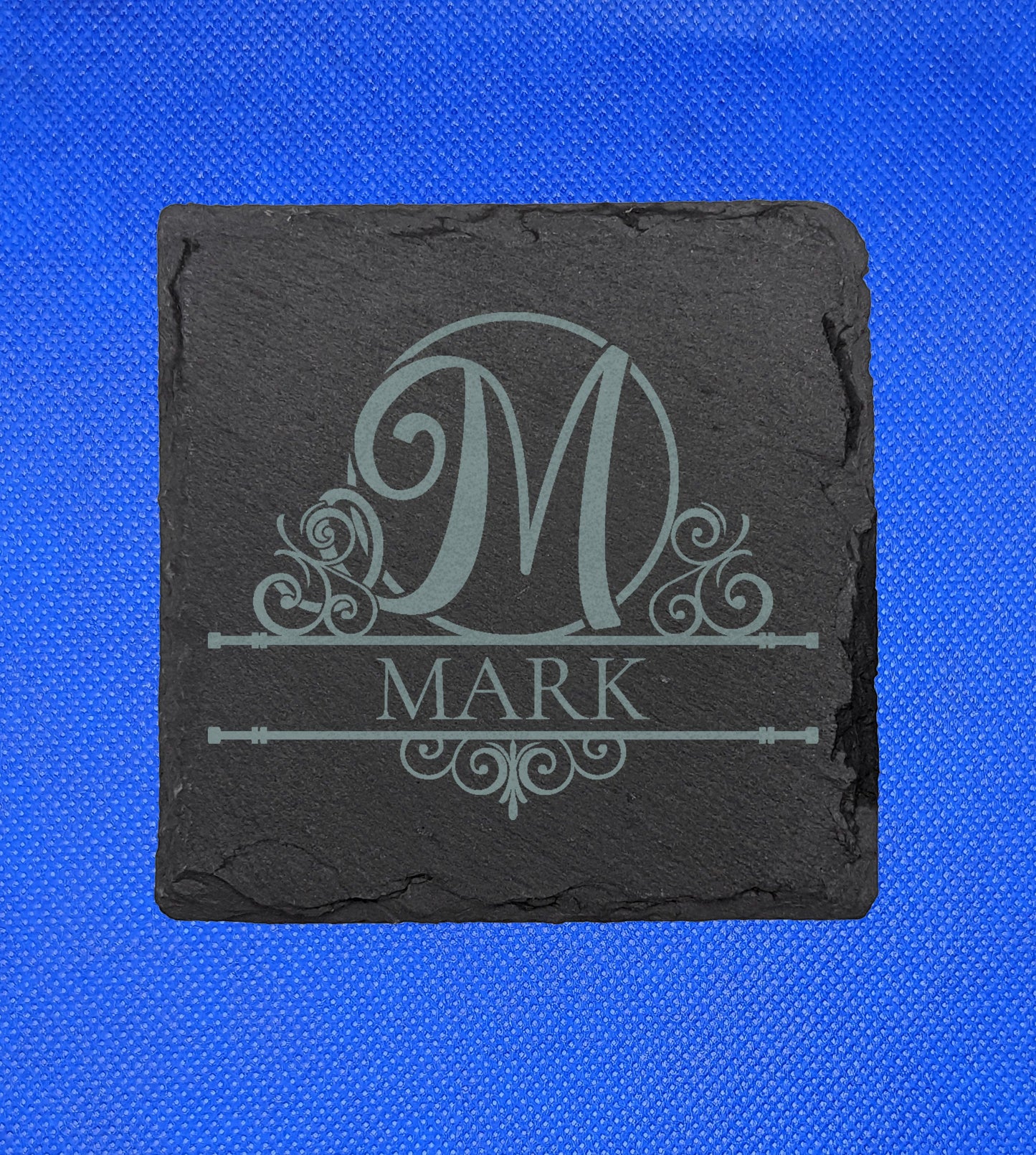 Shows a slate square coaster engraved with the example Initial M in elegant lettering and fancy patterns around it. Under is the name MARK between two horizontal bars with more patterns. It is against a blue background.