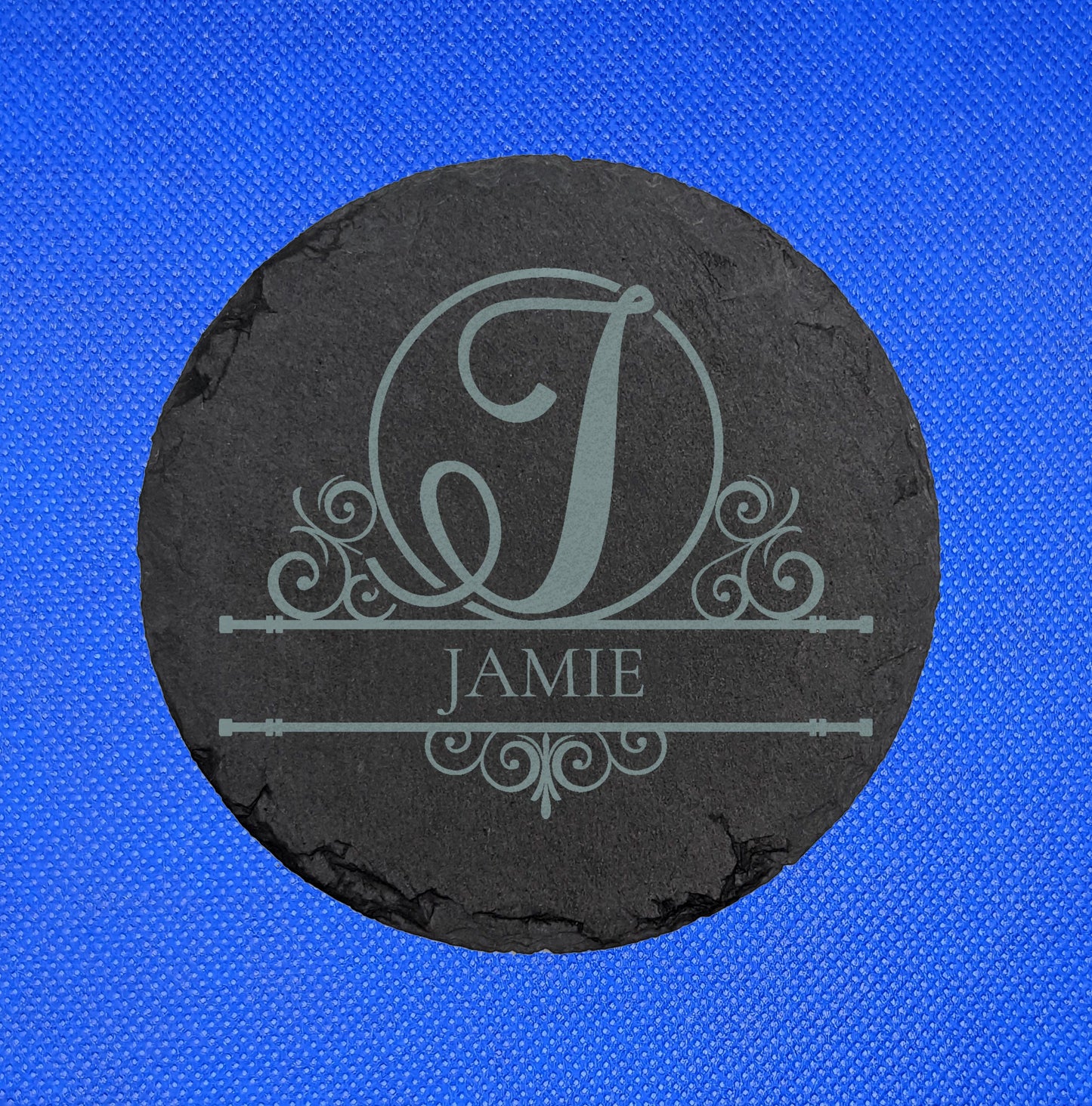 Shows a slate heart coaster engraved with the example Initial J in elegant lettering and fancy patterns around it. Under is the name JAMIE between two horizontal bars with more patterns. It is against a blue background.