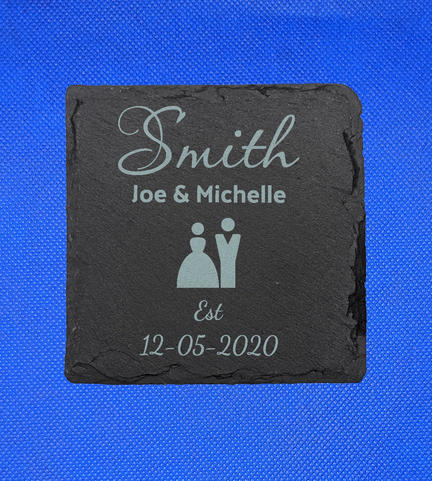 Shows a Wedding Themed square slate coaster 10cm by 10cm. It is engraved with example text saying Smith Joe and Michelle Est 12.05.2020 with the image of man and wife symbols in the middle. It is against a blue background.