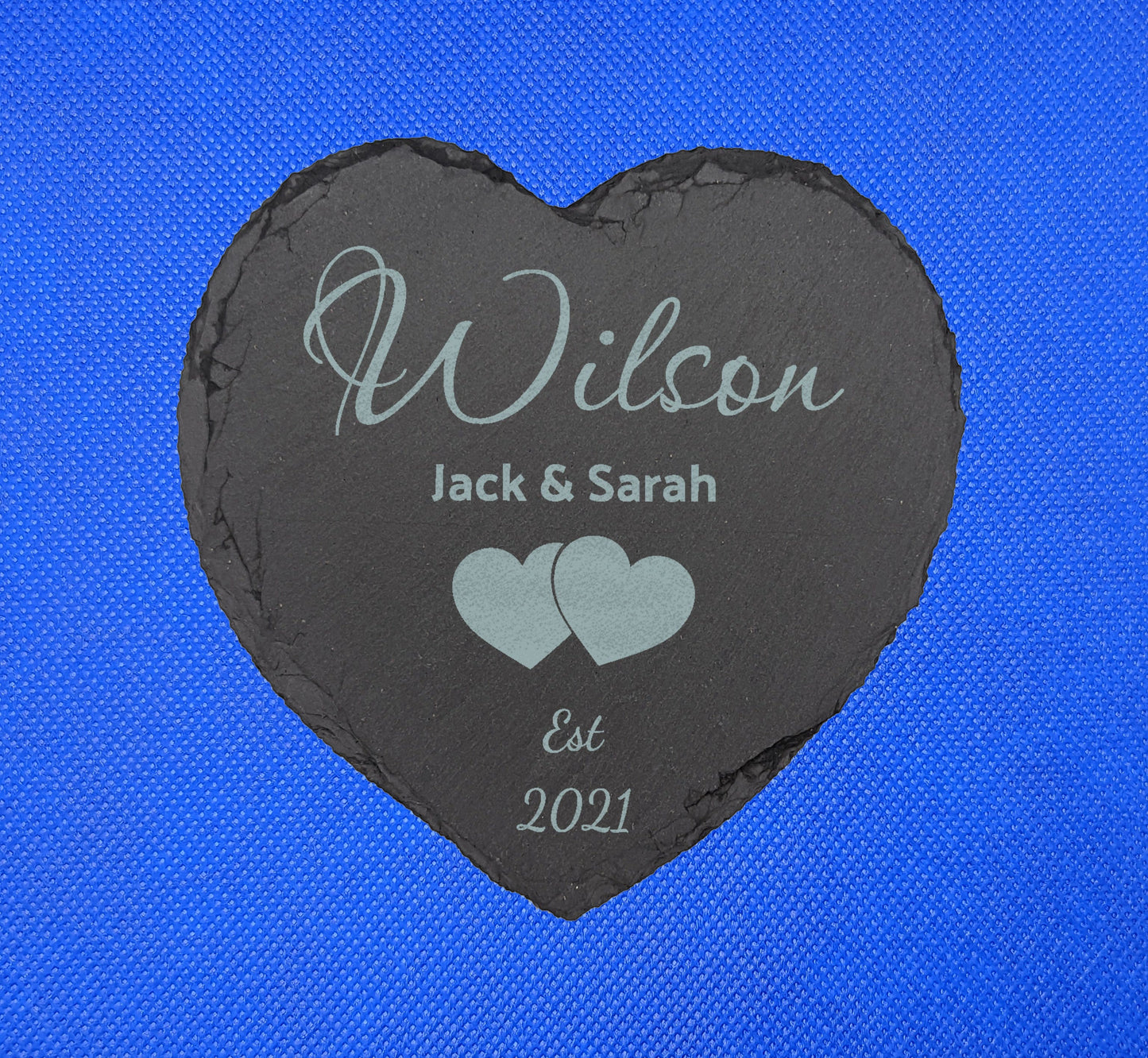 Shows a Wedding Themed heart slate coaster 10cm by 10cm. It is engraved with example text saying Wilson Jack and Sarah Est 2021 with the image of two overlapping hearts in the middle. It is against a blue background.