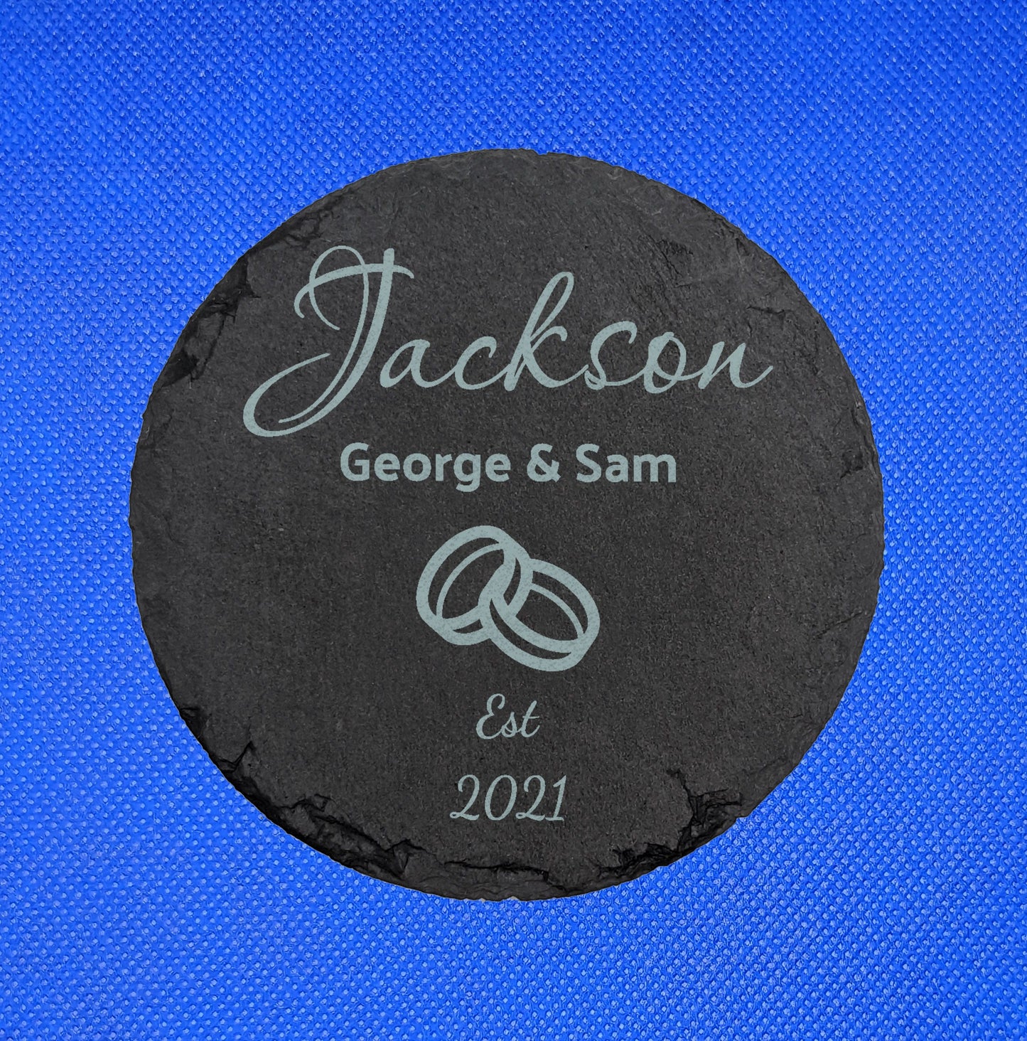 Shows a Wedding Themed slate circle coaster 10cm by 10cm. It is engraved with example text saying Jackson George and Sam Est 2021 with the image of two interlocking rings in the middle. It is against a blue background.