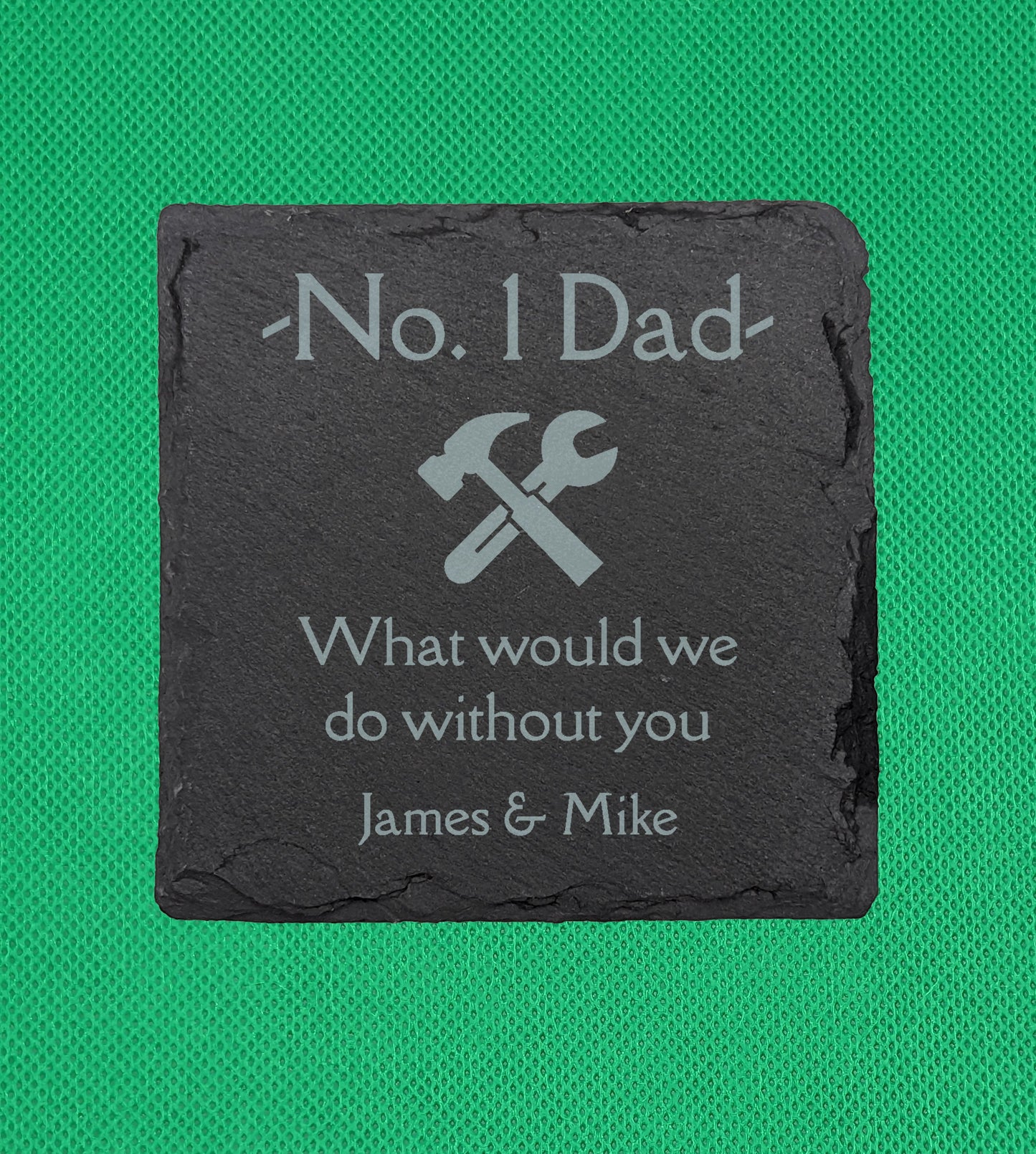 Shows a male themed slate Square coaster 10cm by 10cm. It is engraved with example text saying No. 1 Dad What would we do without you with an image of a wrench and hammer in the middle. It is against a Green background.