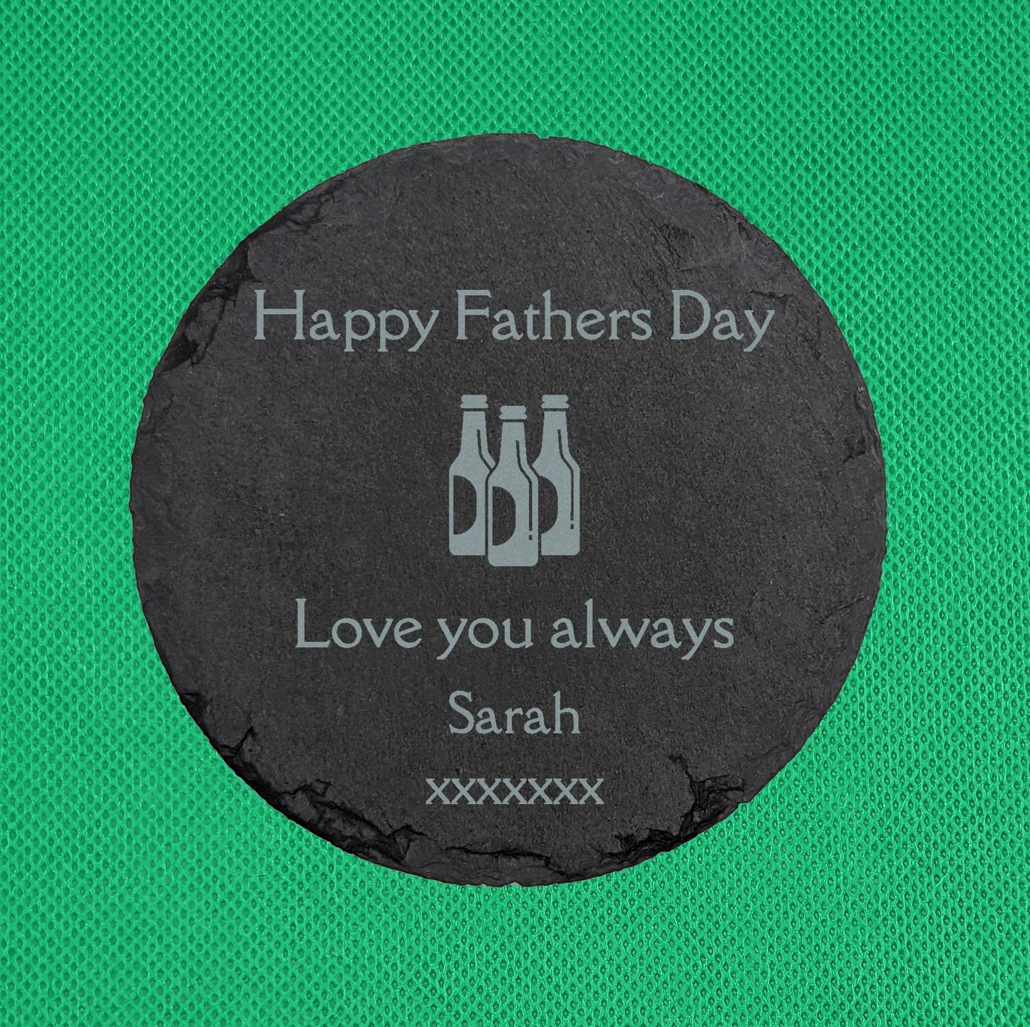 Shows a male themed Slate circle coaster 10cm by 10cm. It is engraved with example text saying Happy Fathers day Love you always Sarah xxxxxxx with an image of Three Beer Bottles in the middle. It is against a Green background.