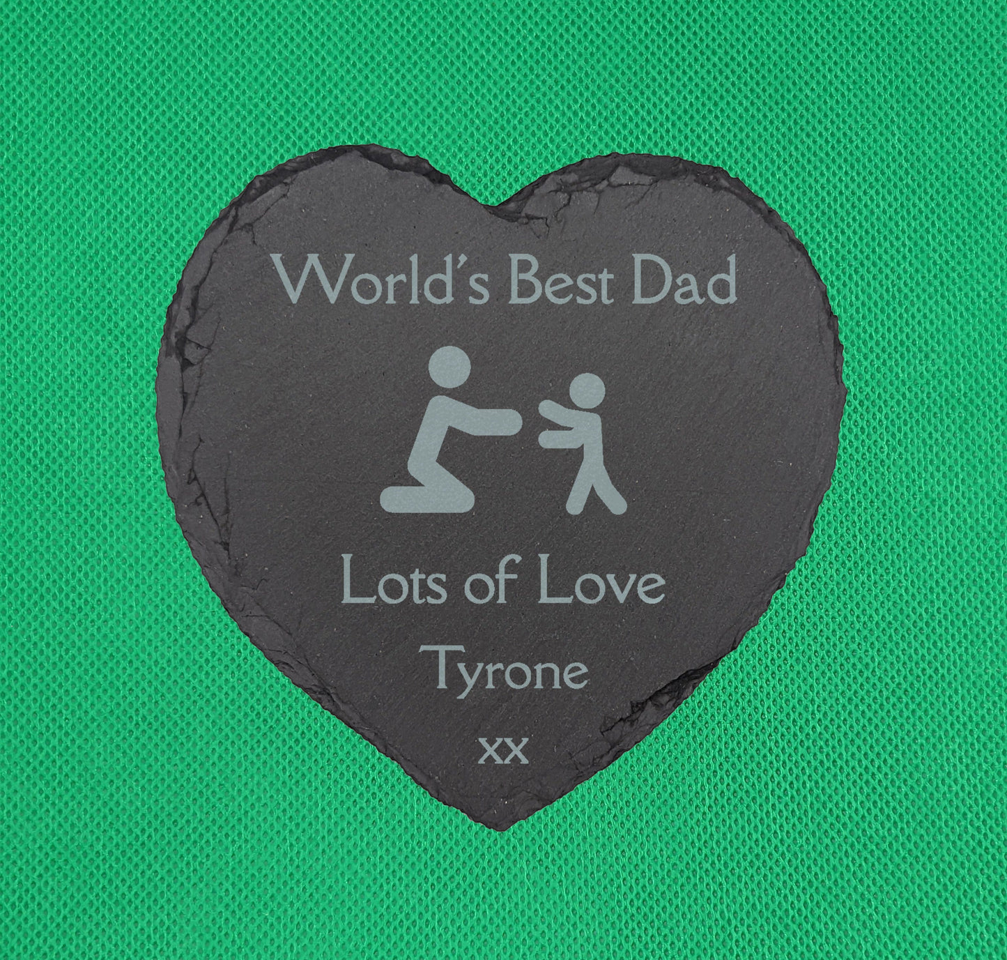 Shows a male themed Slate heart coaster 10cm by 10cm. It is engraved with example text saying Worlds Best Dad Lots of Love Tyrone xx with an image of Stick figure Dad and a son in the middle. It is against a Green background.