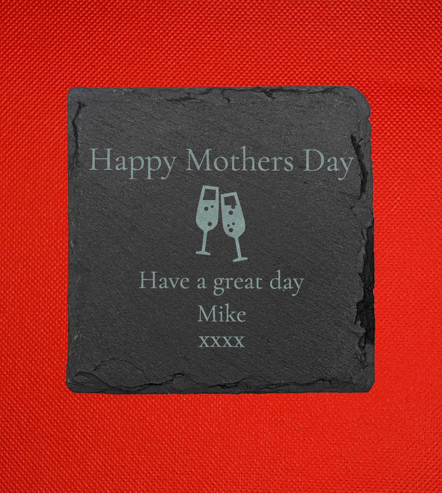 Shows a mum themed square slate coaster 10cm by 10cm. It is engraved with example text saying Happy Mothers Day Have a great day Mike xxxx with an image of Two Champaign Glasses in the middle. It is against a Red background.