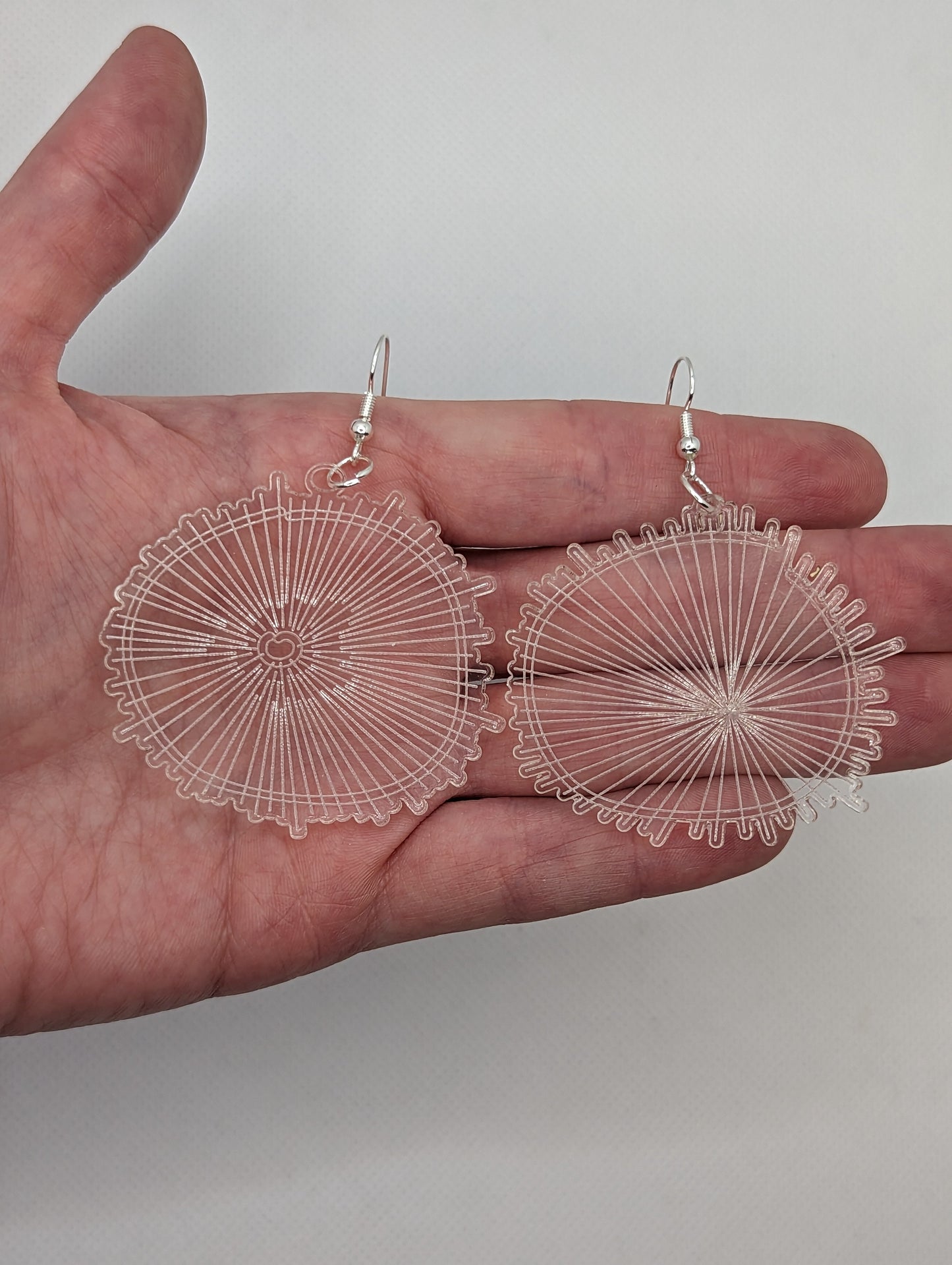 A pair of Clear acrylic earrings with Silver ear wire hook fixings. They are round with a web engraved on them which also looks like a wood slice pattern. They are against a hand to show the size of three finger widths in length and same again wide.