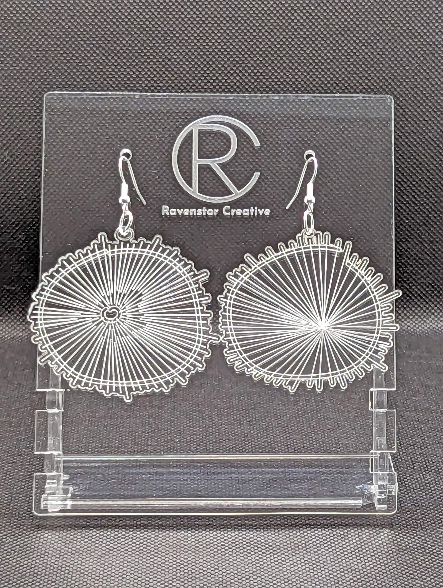 A pair of Clear acrylic earrings with Silver ear wire hook fixings. They are round with a web engraved on them which also looks like a wood slice pattern. They are against a black background on a clear stand with the Ravenstor Creative logo on it.