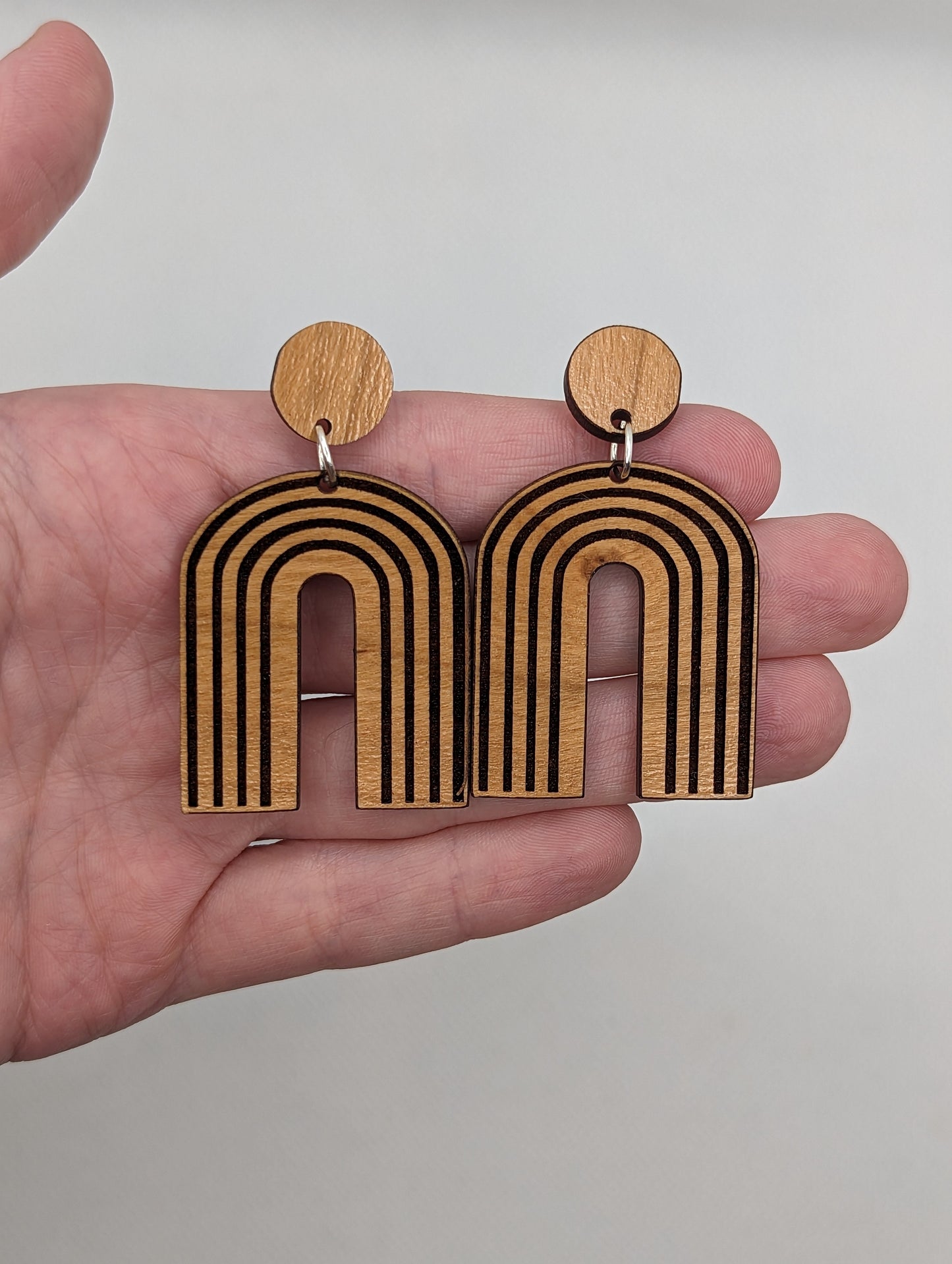 A pair of arch earrings dangling under a circle with stud fixings. Engraved are lines like a wooden rainbow 80s retro style. They are made from cherry wood. They are against a hand to show the size of three finger widths in length and two wide